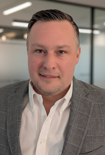 Professional headshot of a man with short dark hair, wearing a gray checked blazer and white shirt, standing in an office with glass walls.
