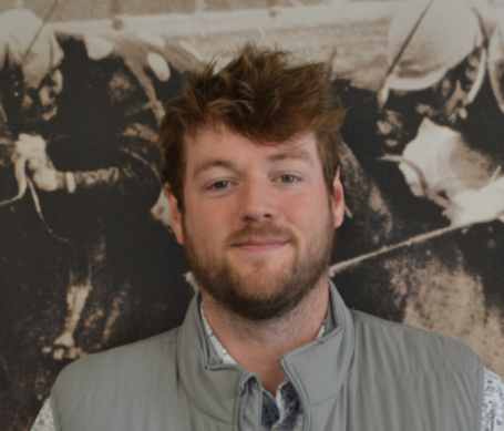 A man with light skin, brown curly hair, and a beard, smiling, wearing a gray vest over a patterned shirt, standing in front of a black-and-white historical photo background.