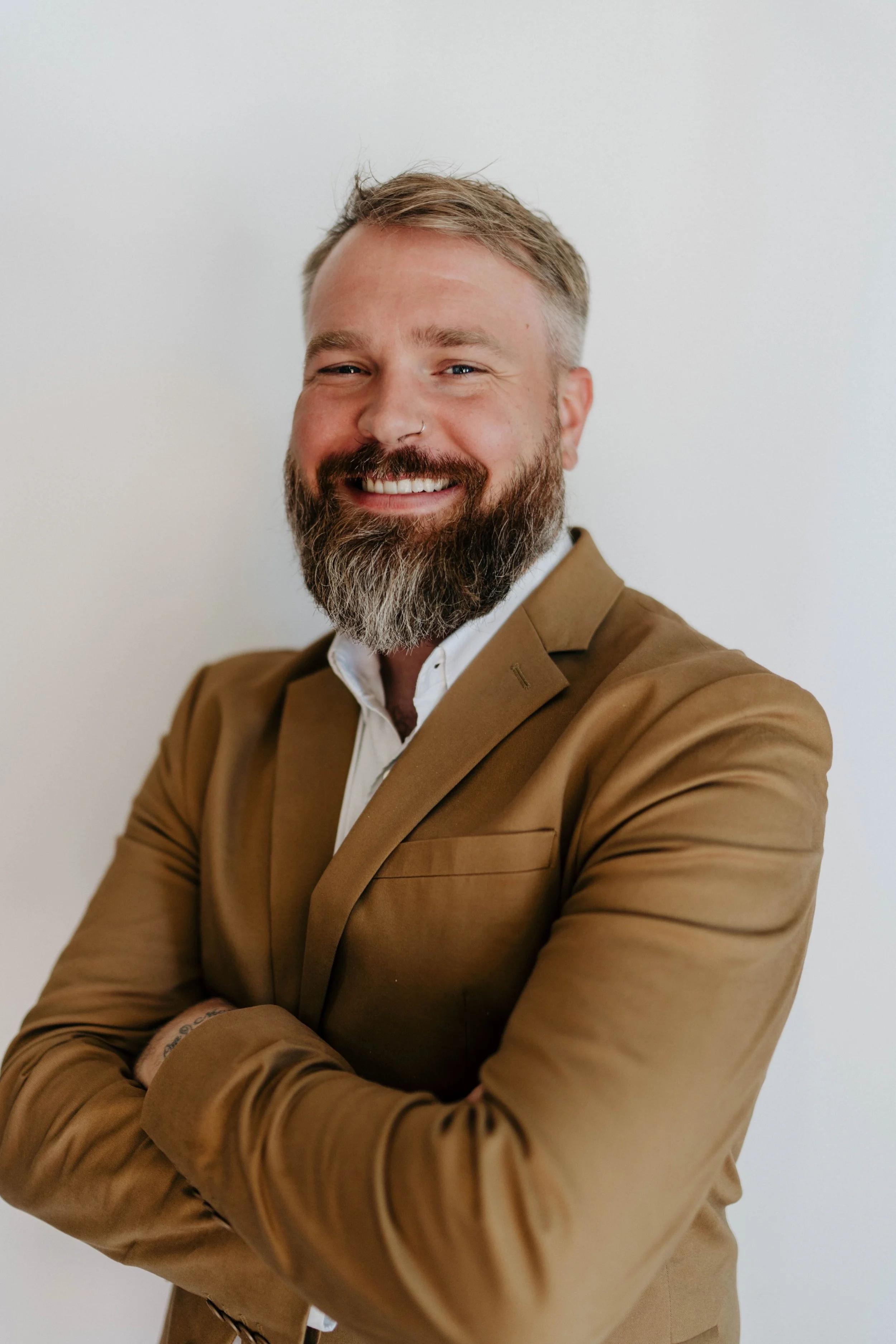 A smiling man with a beard and light-colored hair, wearing a tan suit and white shirt, standing against a plain white background.