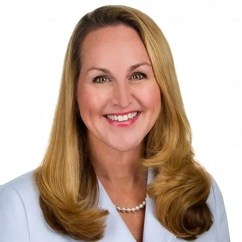 Portrait of a smiling woman with blonde hair wearing a light-colored blazer and pearl necklace.