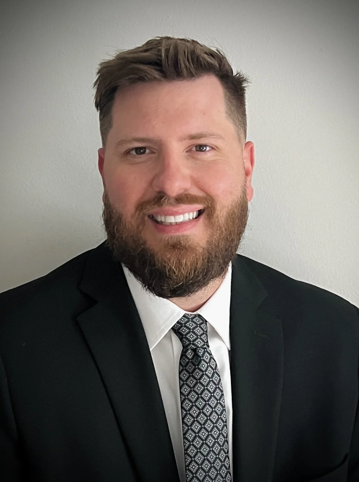A man with a beard and styled hair wearing a black suit, white shirt, and patterned tie, smiling against a plain light gray background.