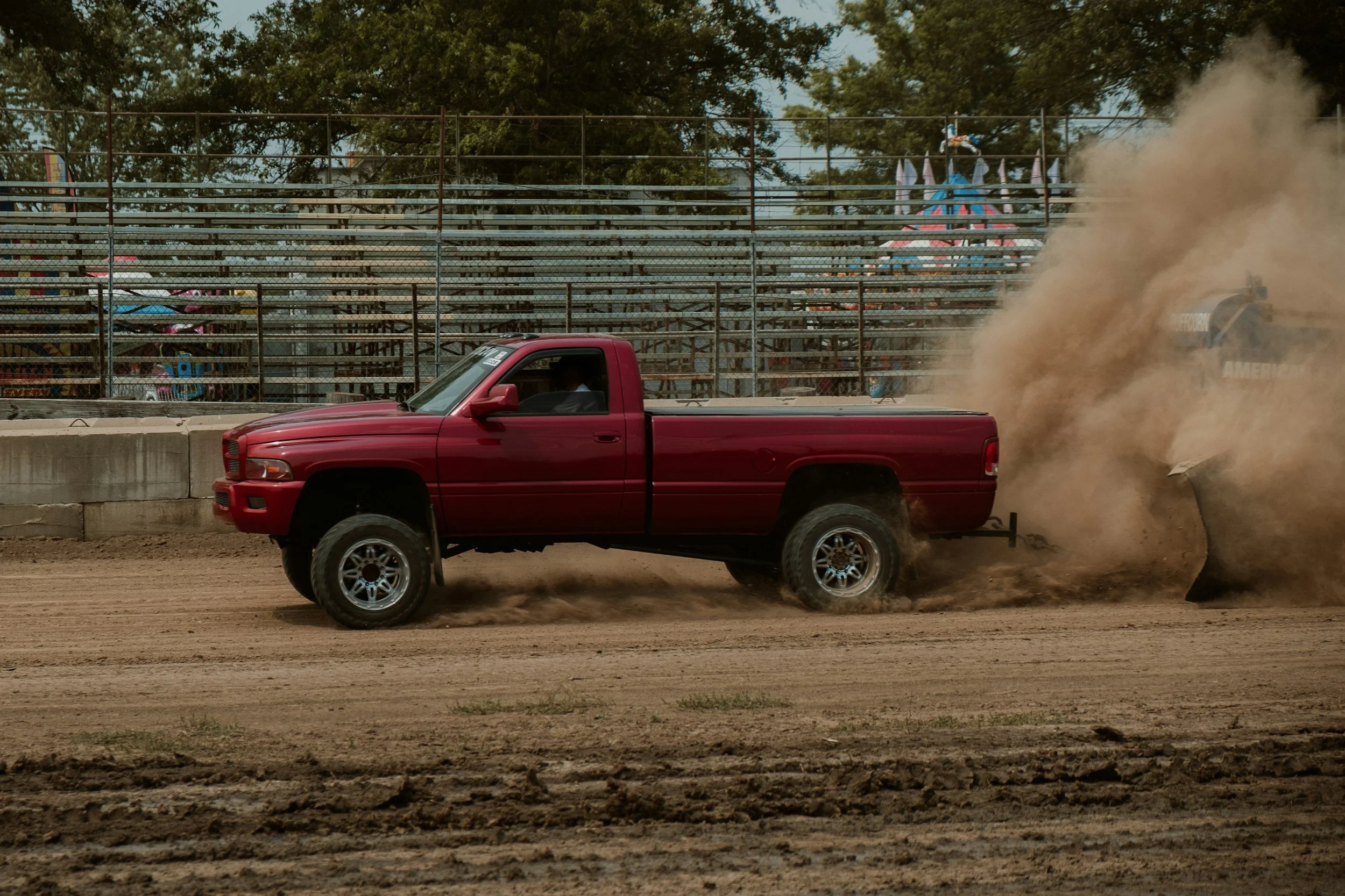 Red pickup truck racing on a dirt track, kicking up dust, with empty bleachers and trees in the background.