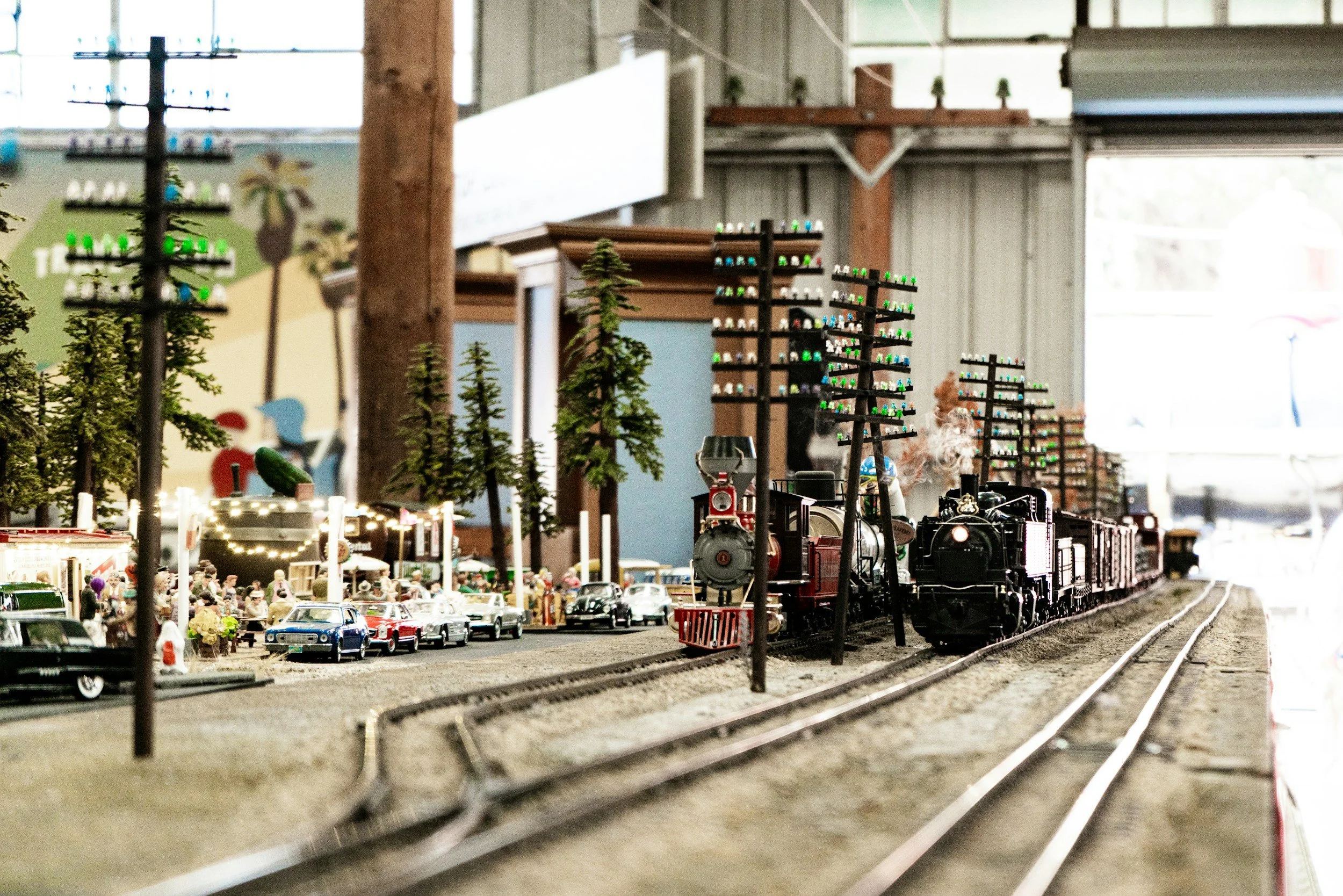 A miniature train set display featuring a black steam locomotive pulling cars along tracks, with small trees, model buildings, and toy cars, in an indoor setting.