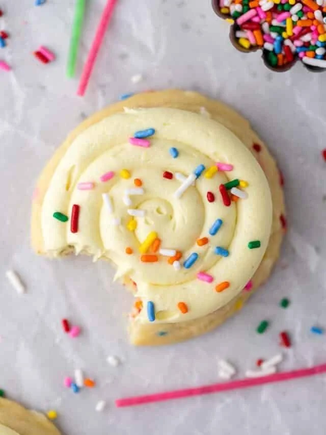 A partially eaten vanilla rolled cake with white frosting and colorful sprinkles on top, on a white surface with scattered sprinkles, and a small dish of additional sprinkles in the background.