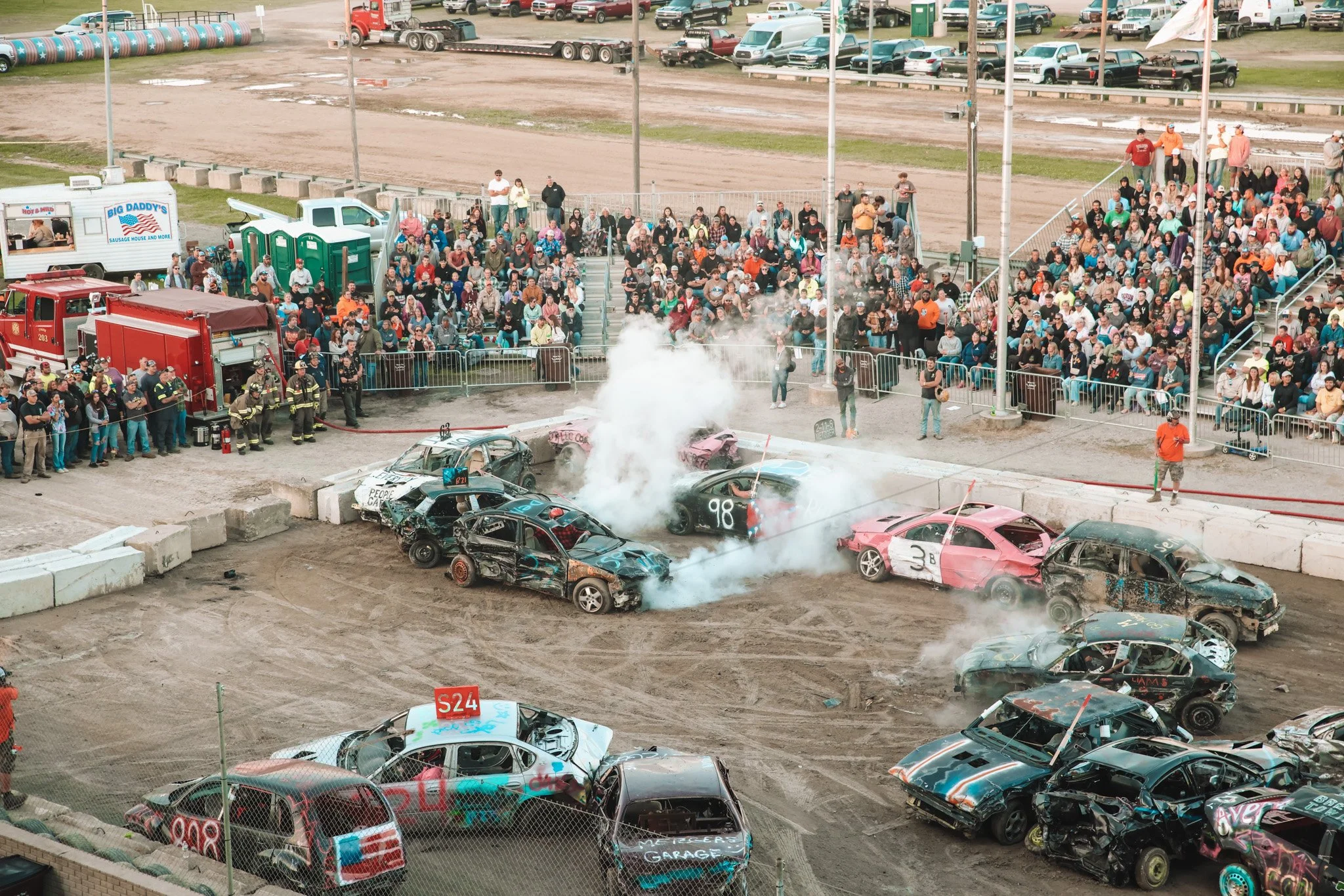 Race car crash with two damaged cars colliding on dirt track, debris flying, in front of empty green grandstands.
