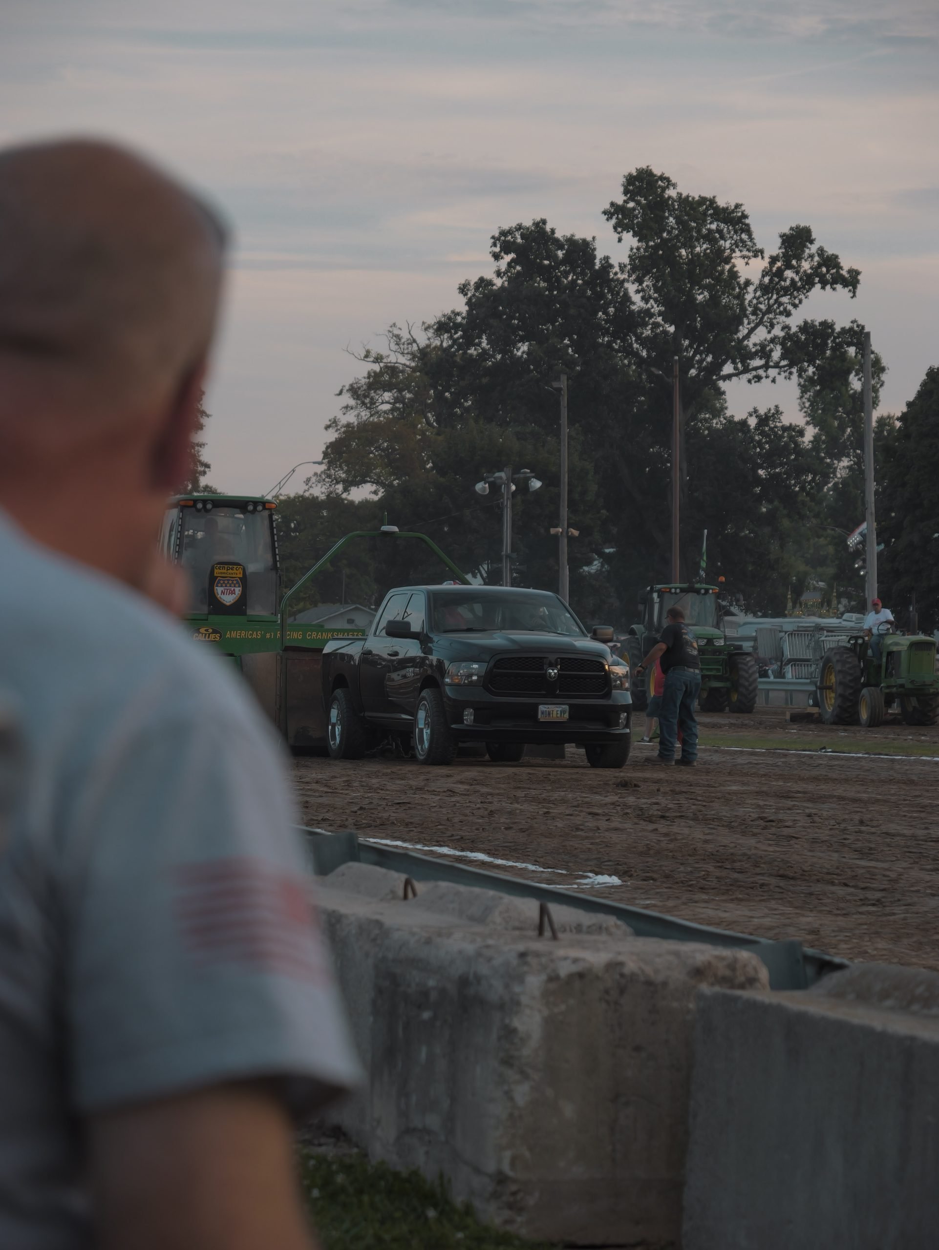 A black pickup truck on a dirt track with agricultural machinery and farm equipment in the background, and a blurred person in the foreground.