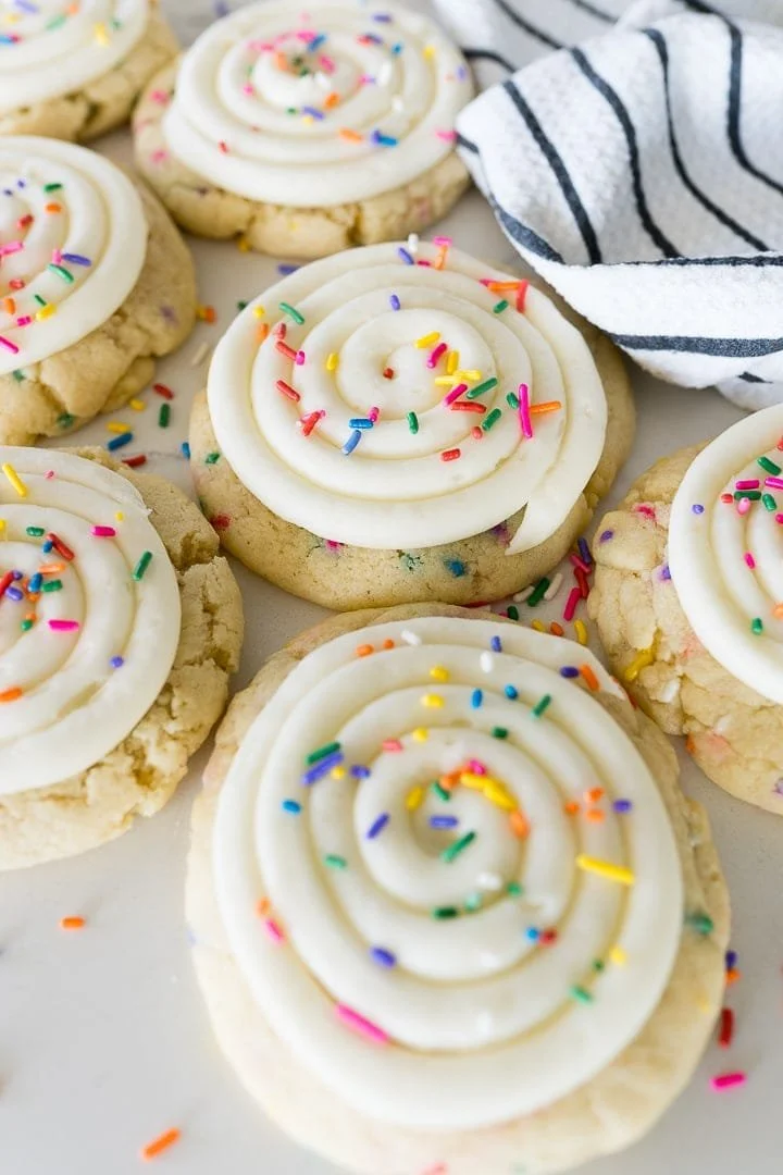 Cookies decorated with white icing in a swirl design and colorful sprinkles, with a black and white striped cloth nearby.