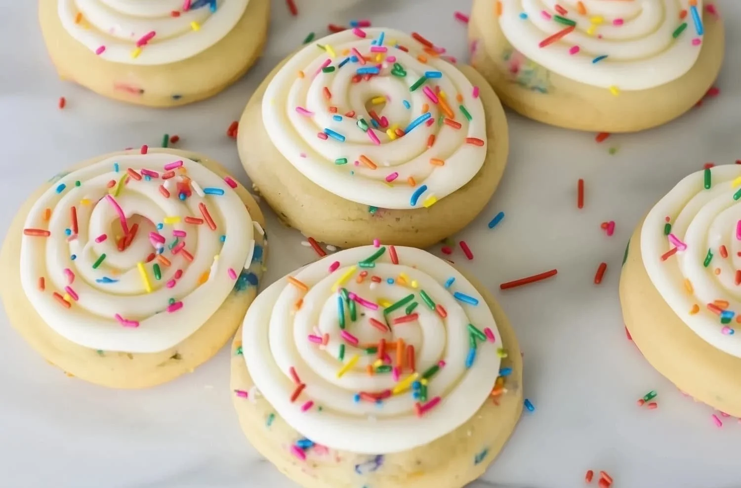 Close-up of several vanilla cookies topped with white frosting and colorful rainbow sprinkles.
