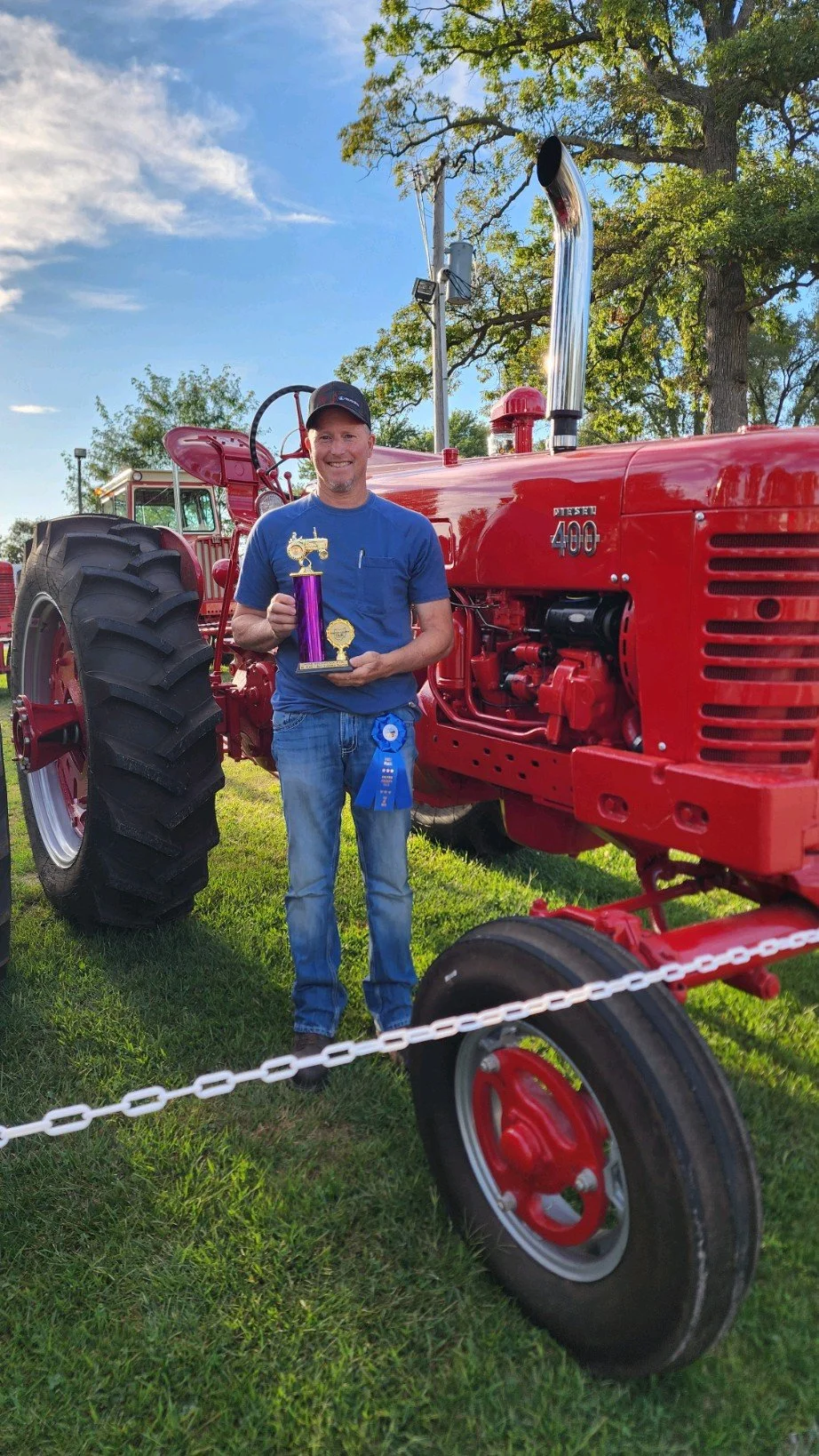 Man smiling and holding a trophy, standing next to a vintage red tractor outdoors on grass, with trees and blue sky in the background.