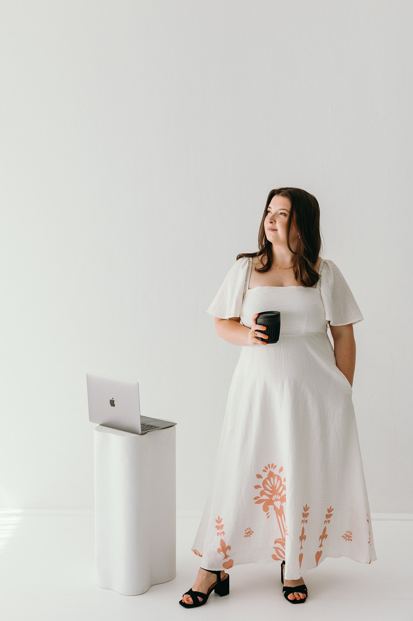 Lauren Hunter of Unravel standing confidently in a bright studio, wearing a white floral-print dress and holding a coffee cup, with a MacBook on a white pedestal beside her, representing the Unravel and Elevate service.