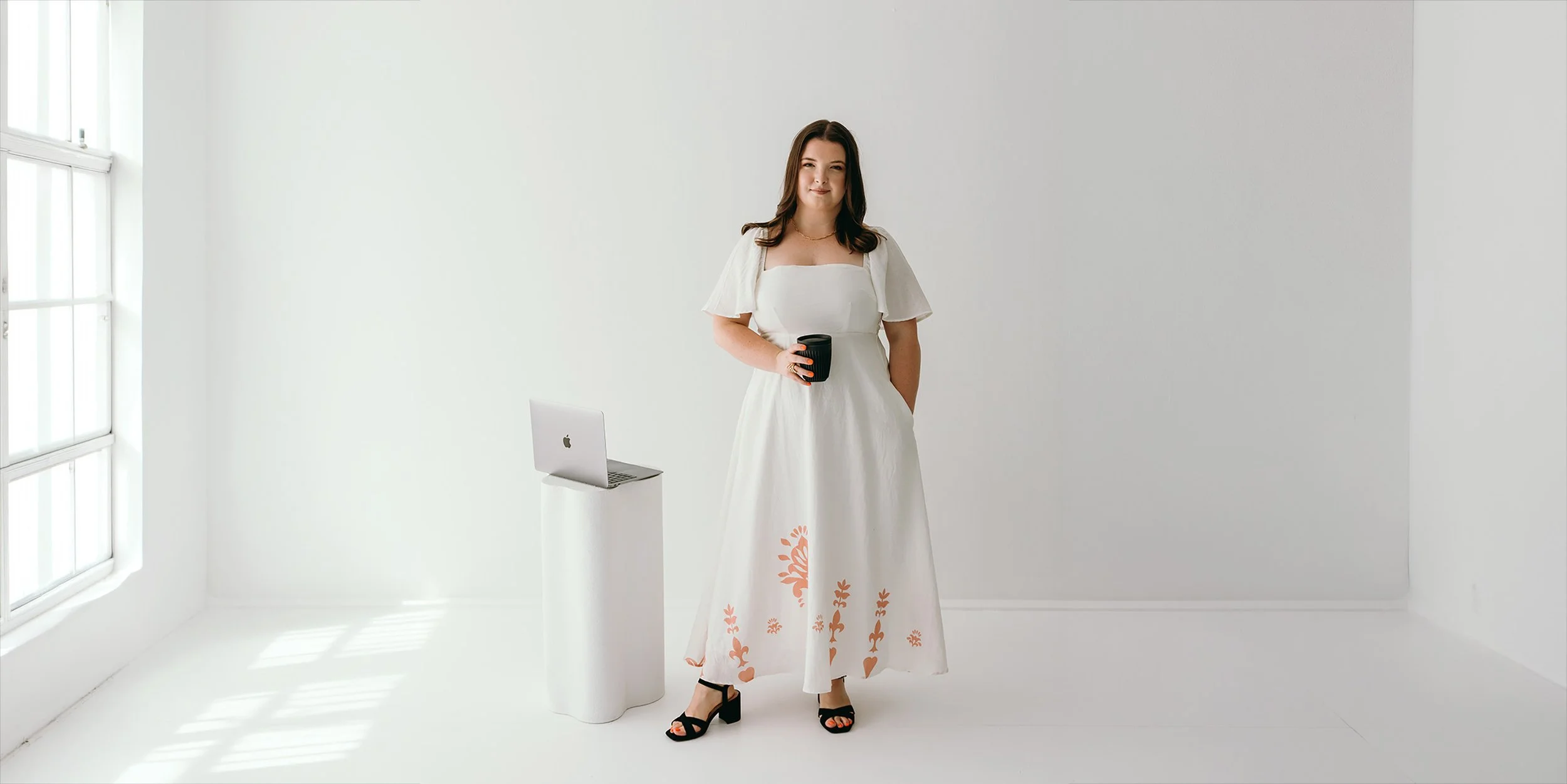 Lauren Hunter of Unravel standing confidently in a bright studio, wearing a white floral-print dress and holding a coffee cup, with a MacBook on a white pedestal beside her, representing the Unravel and Elevate service.