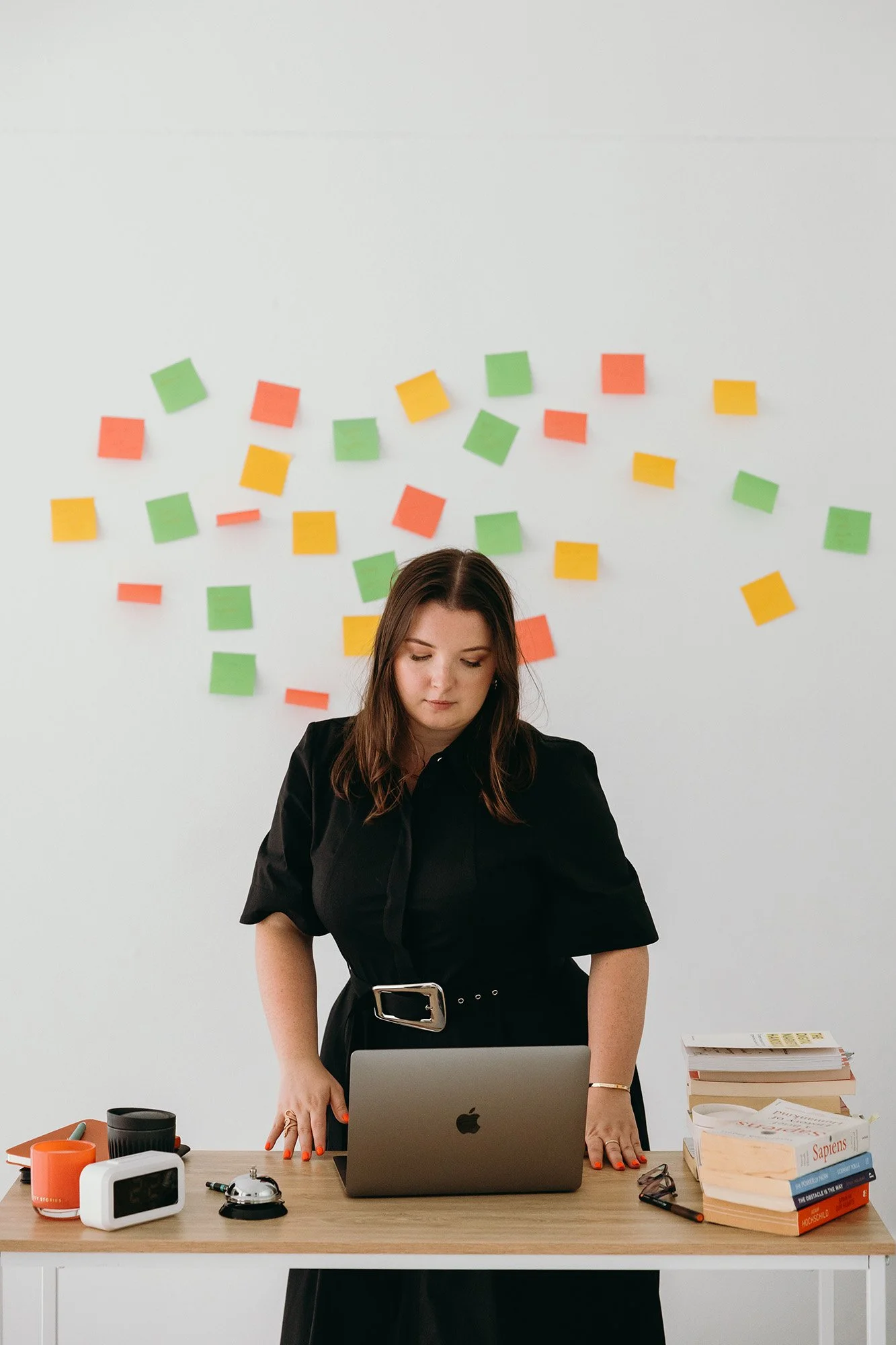 Lauren, Founder of Unravel, is standing at a desk with a laptop, surrounded by books, a clock, and office supplies, with colorrful sticky notes on the white wall behind her.