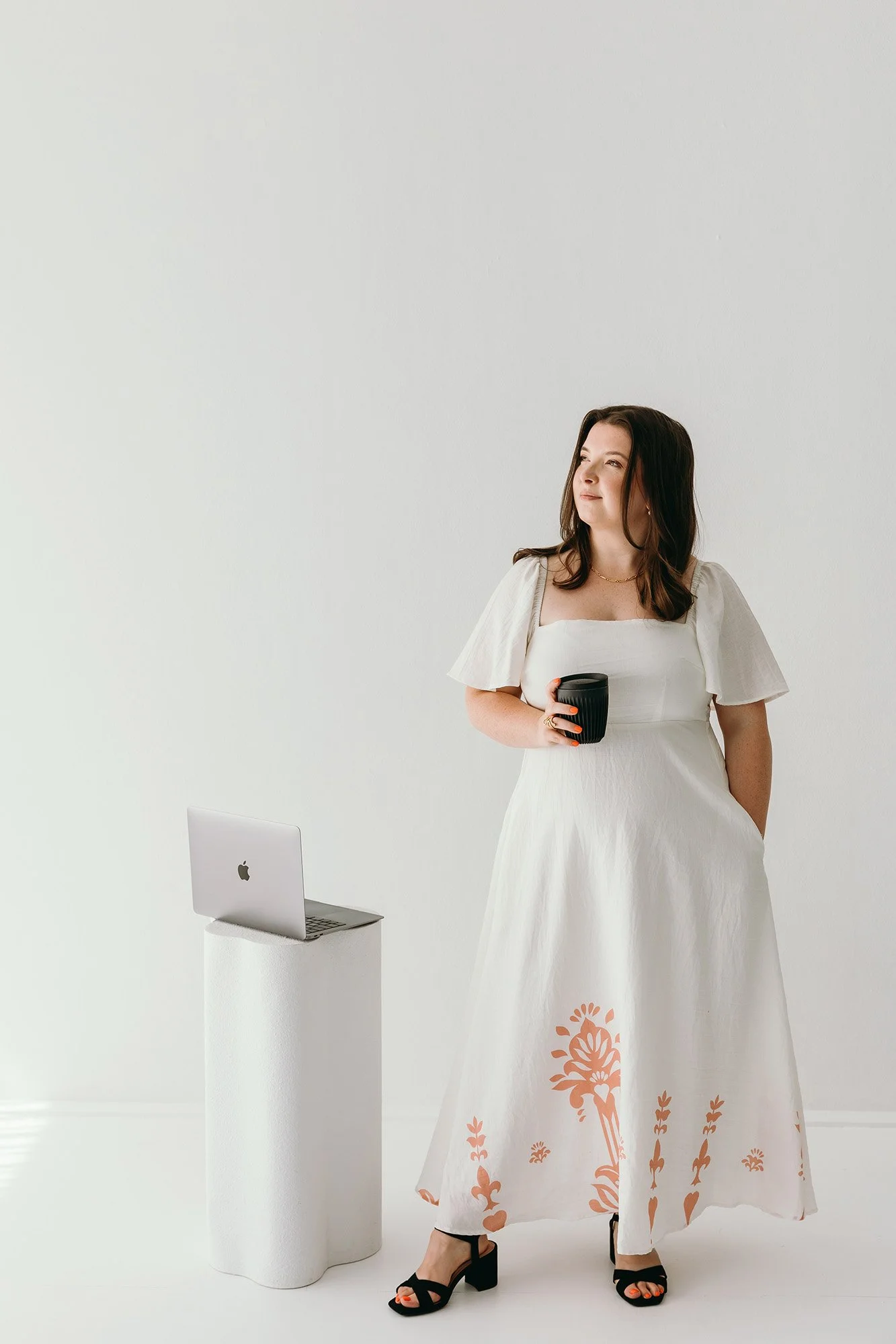 Lauren Hunter of Unravel standing confidently in a bright studio, wearing a white floral-print dress and holding a coffee cup, with a MacBook on a white pedestal beside her, representing the Unravel and Elevate service.