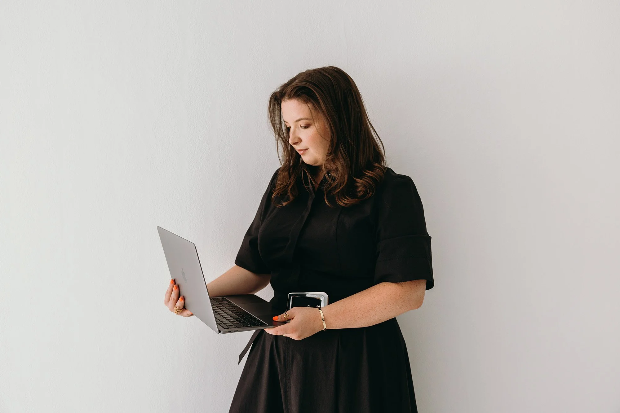 A woman in a black dress holds an open laptop and phone while reviewing her work, representing Unravel's hands-on approach to setting up client systems, emails, and funnels.