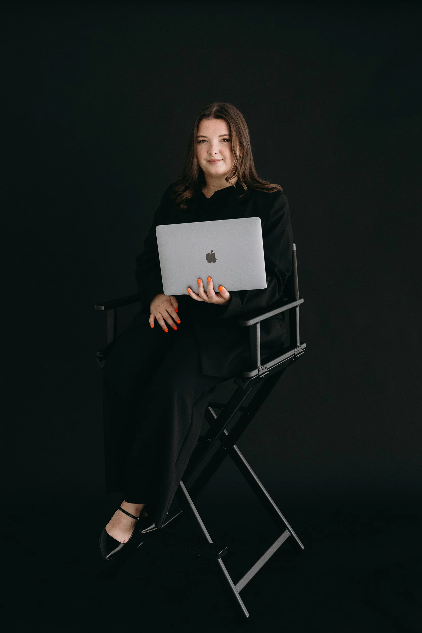 Lauren, Founder of Unravel is wearing a black blazer and black trousers, seated on a director's chair against a black background. She is holding a silver MacBook laptop and looking at the camera with a slight smile.
