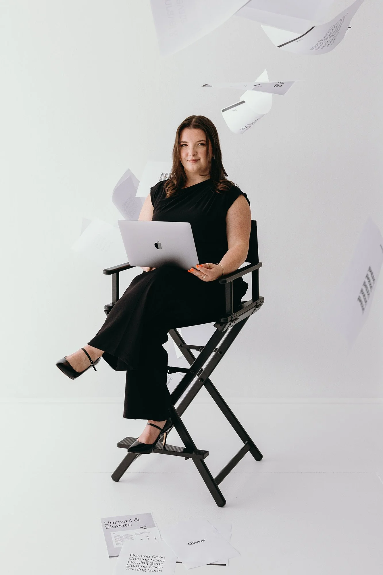 Lauren Hunter of Unravel seated confidently in a director's chair with a MacBook, surrounded by floating and scattered Unravel branded documents, capturing the transformation from business chaos to clarity.