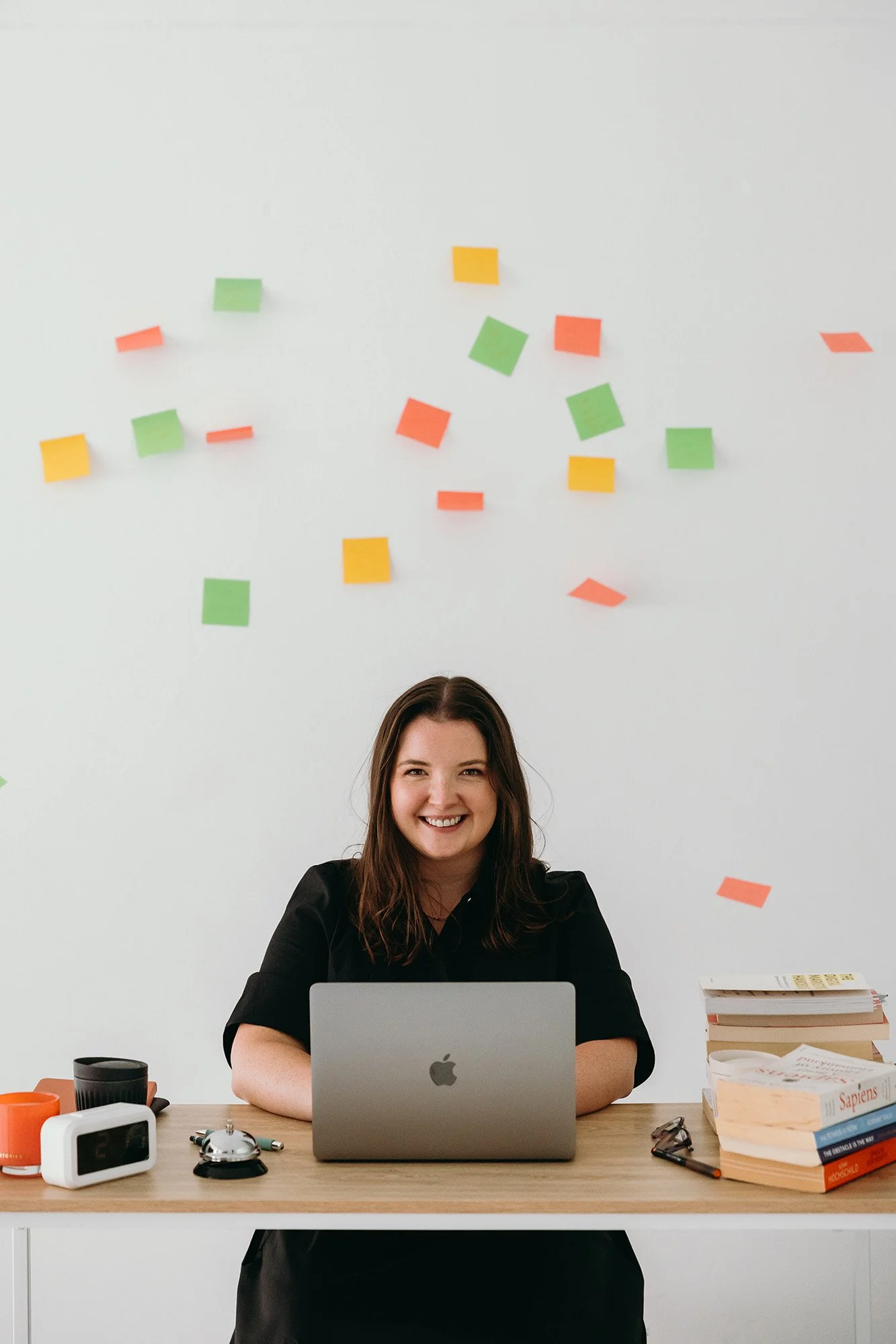 Lauren Hunter, founder of Unravel, smiling warmly at her desk with a MacBook, stack of books, and coffee cups, with colourful sticky notes arranged on the wall behind her.