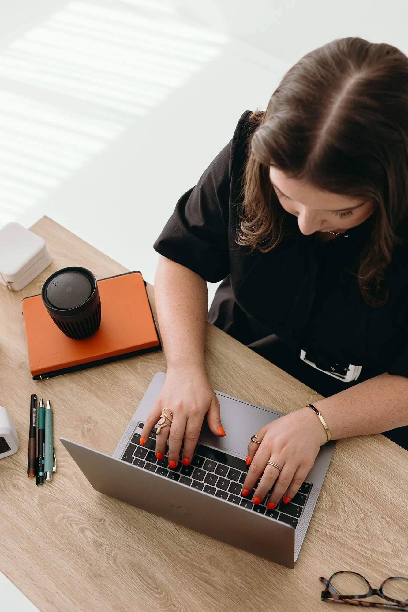 Overhead view of Lauren from Unravel typing on a laptop at a wooden desk, with an orange notebook, coffee cup, pens, and glasses nearby, representing Unravel's 1 to 1 support.