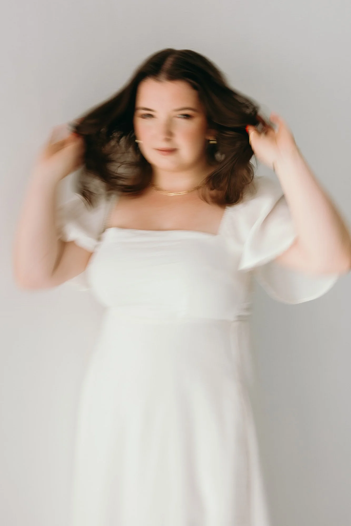 Blurred image of Lauren, Founder of Unravel, in a white dress, standing against a plain background, adjusting her hair.
