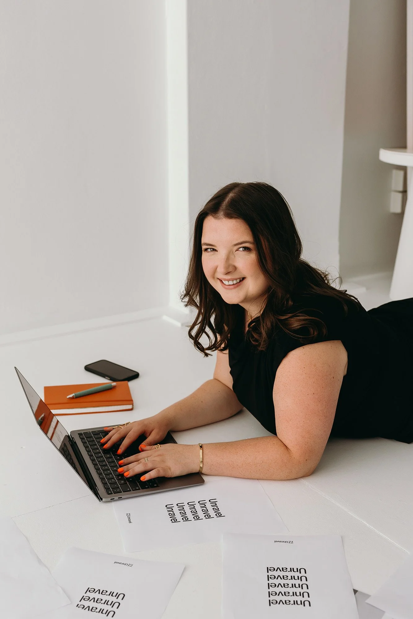 Lauren, Founder of Unravel, is lying on the floor using a laptop, surrounded by printed sheets and notebooks.