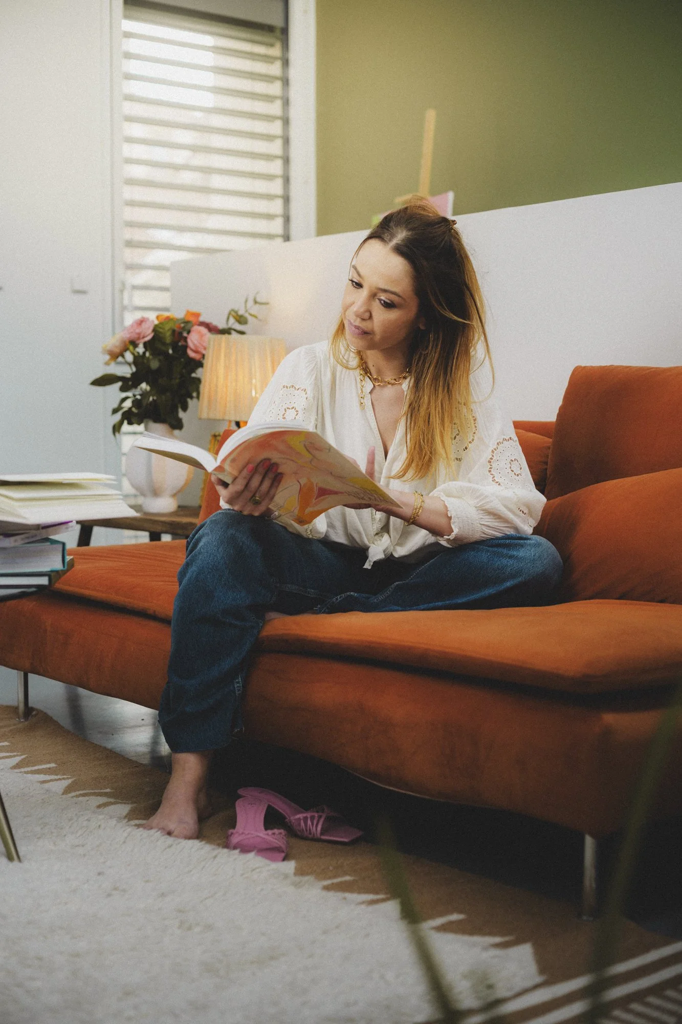 Une femme assise sur un canapé orange, lisant un livre dans un salon avec une lampe de table, un vase de fleurs roses, et une fenêtre avec des volets en arrière-plan.