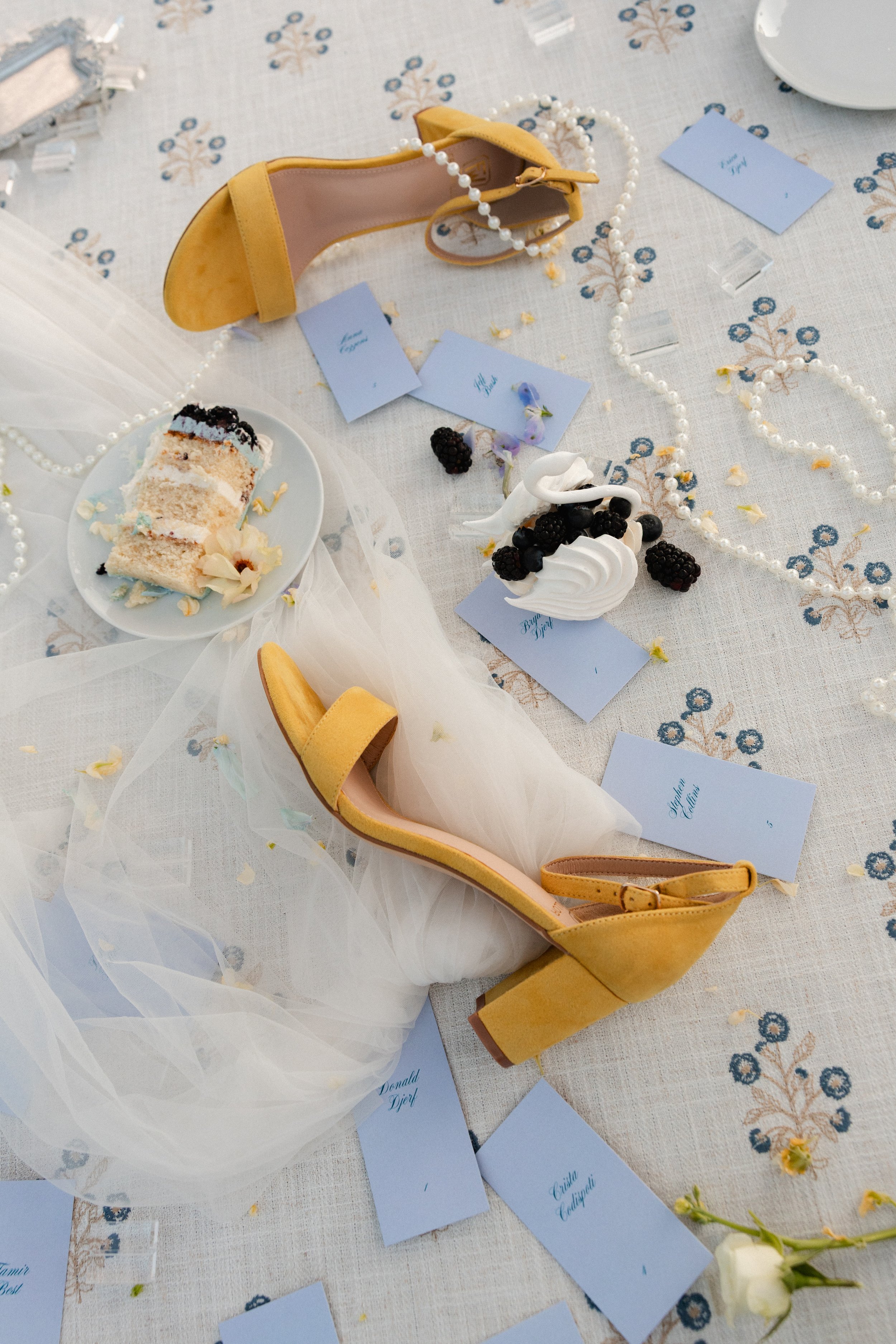 Yellow high-heeled shoes on a decorated table with pearls, blue place cards, a slice of cake with berries, and scattered flower petals.
