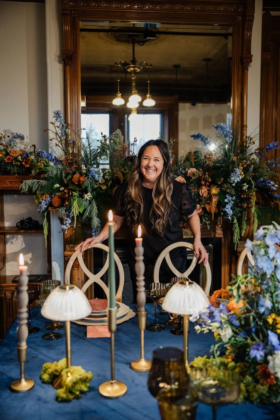 A woman standing behind a dining table decorated with candles and flowers, smiling indoors with a large mirror and chandelier in the background.