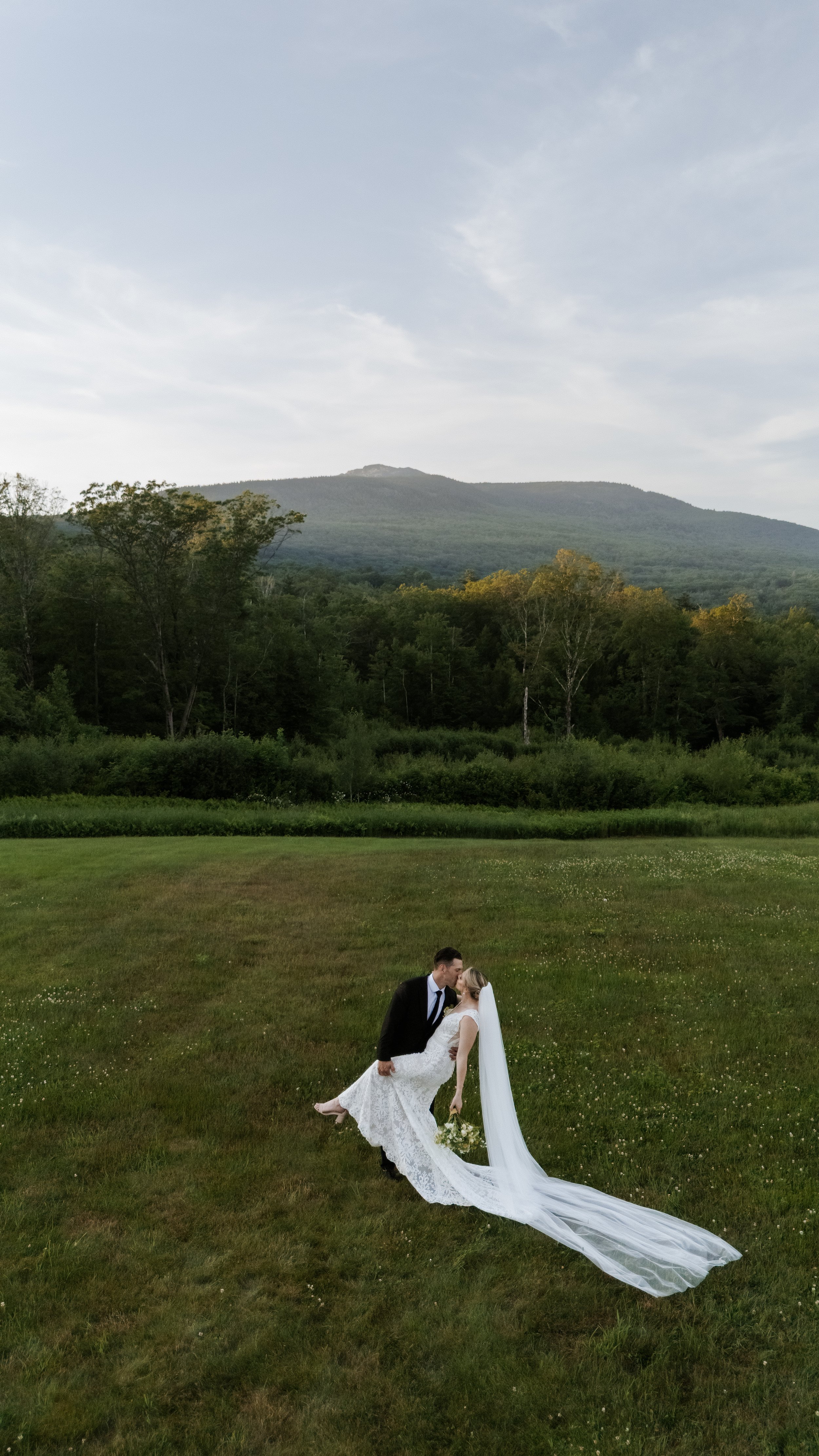 A bride and groom dancing in a field with mountains and trees in the background.