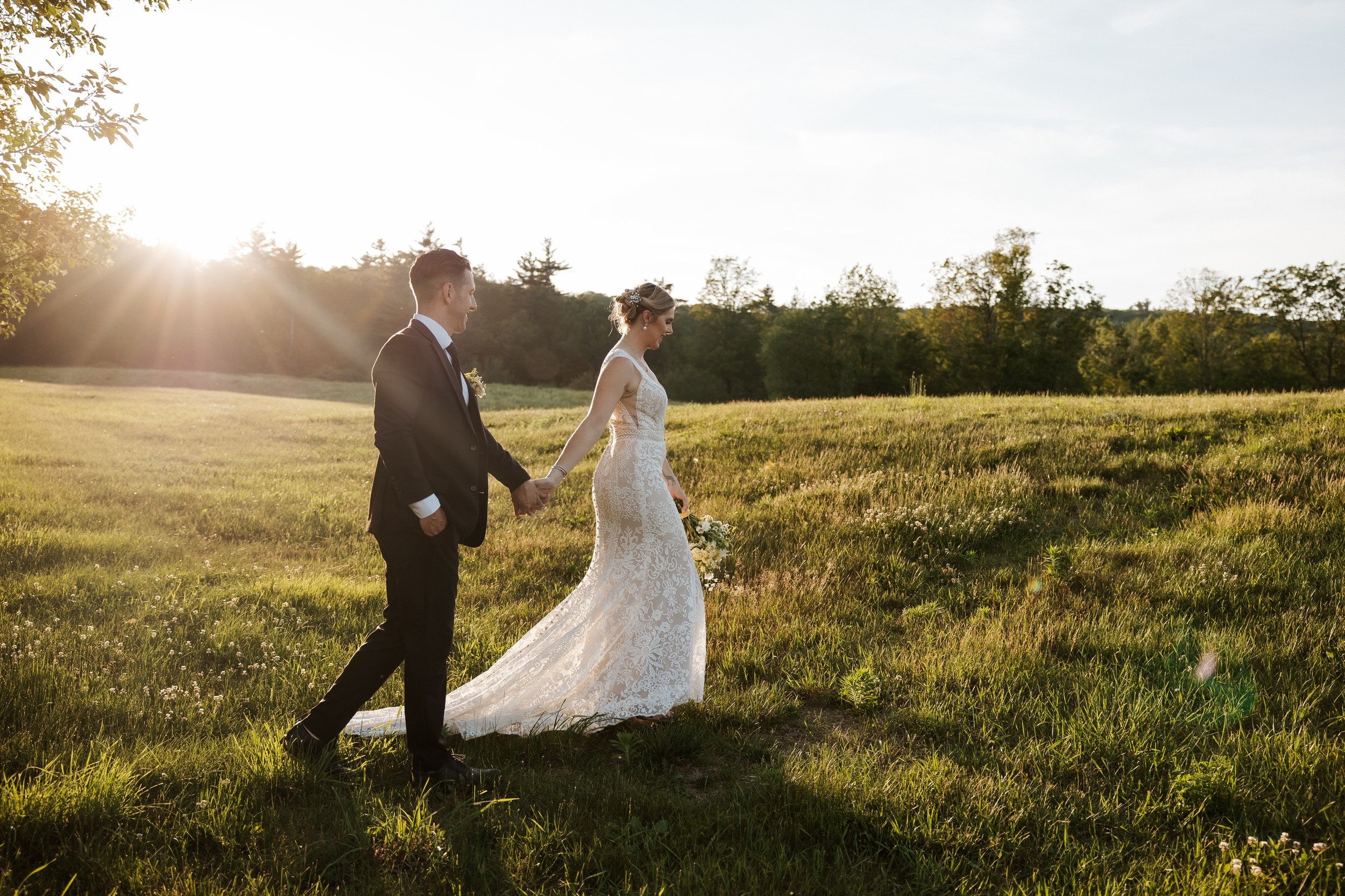 A bride and groom holding hands and walking through a grassy field at sunset, with trees in the background and the sun shining brightly.