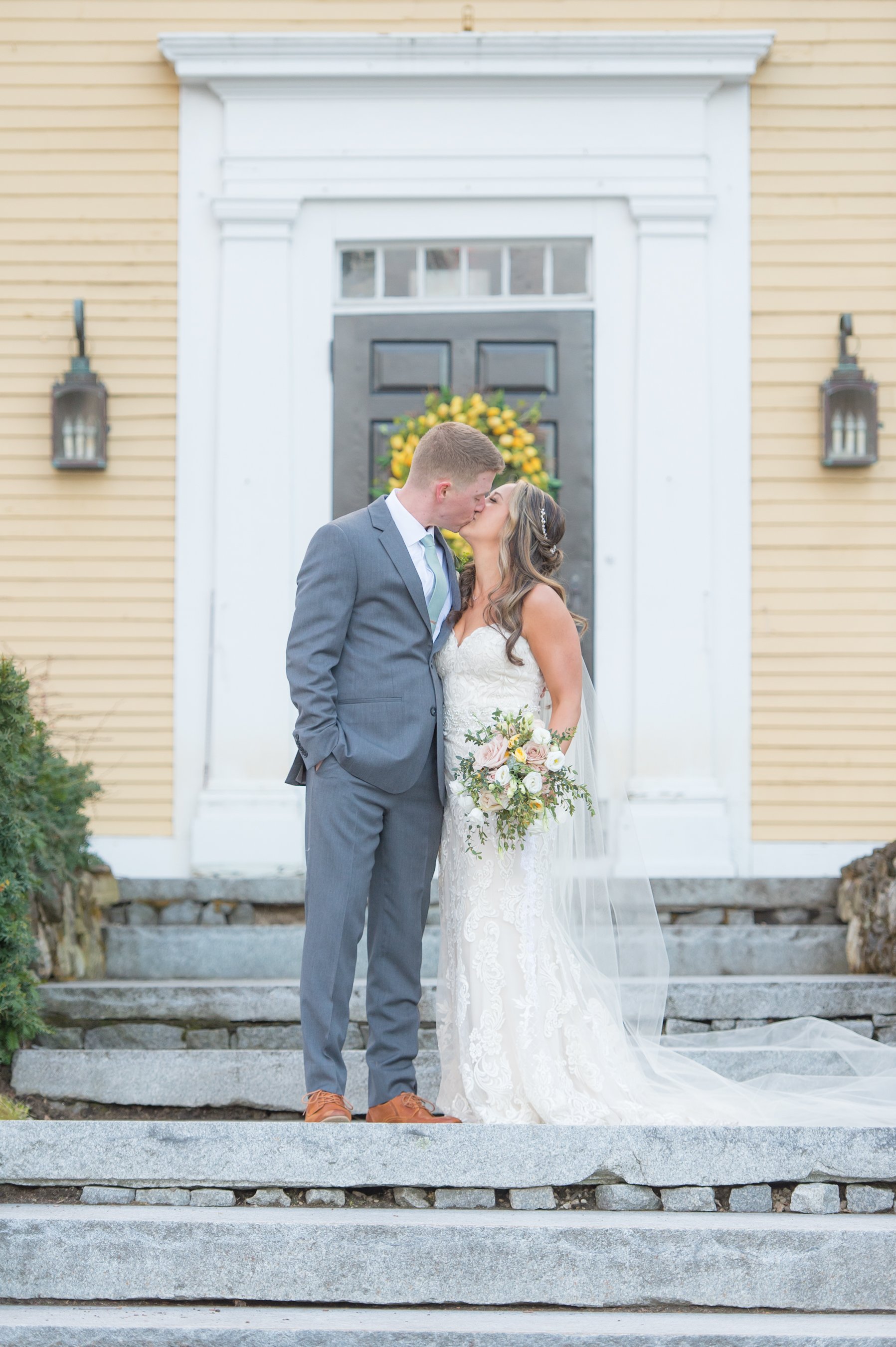 A newlywed couple sharing a kiss on the steps of a house, with the bride holding a bouquet of flowers and wearing a lace wedding dress, and the groom in a gray suit.