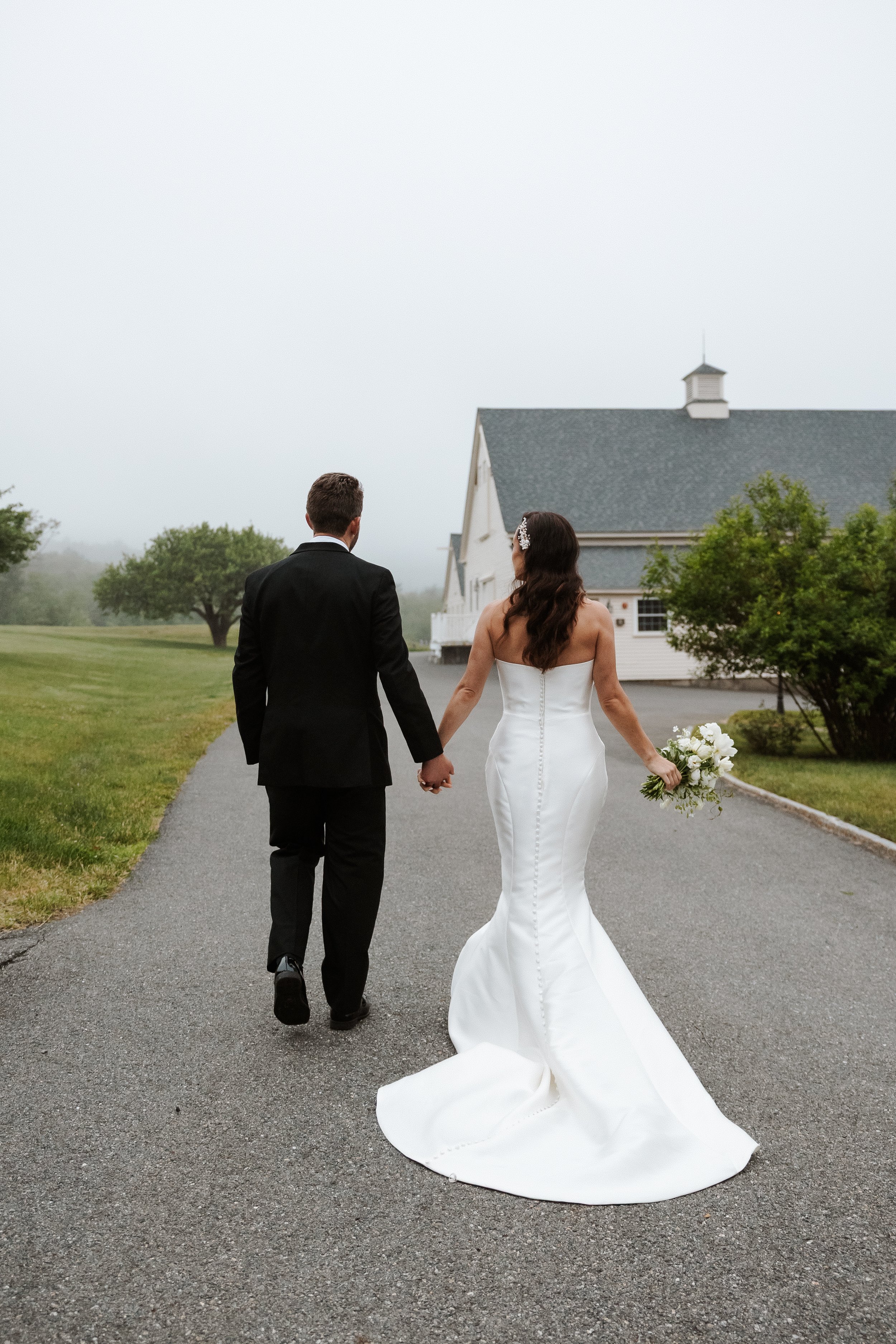 Bride and groom walking hand in hand on a paved path near a white wedding venue, with misty weather and green grass on either side.