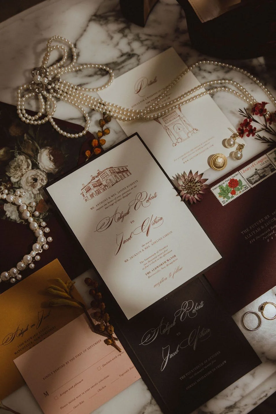 Wedding invitations, pearl necklaces, rings, flowers, and a postage stamp on a marble surface.