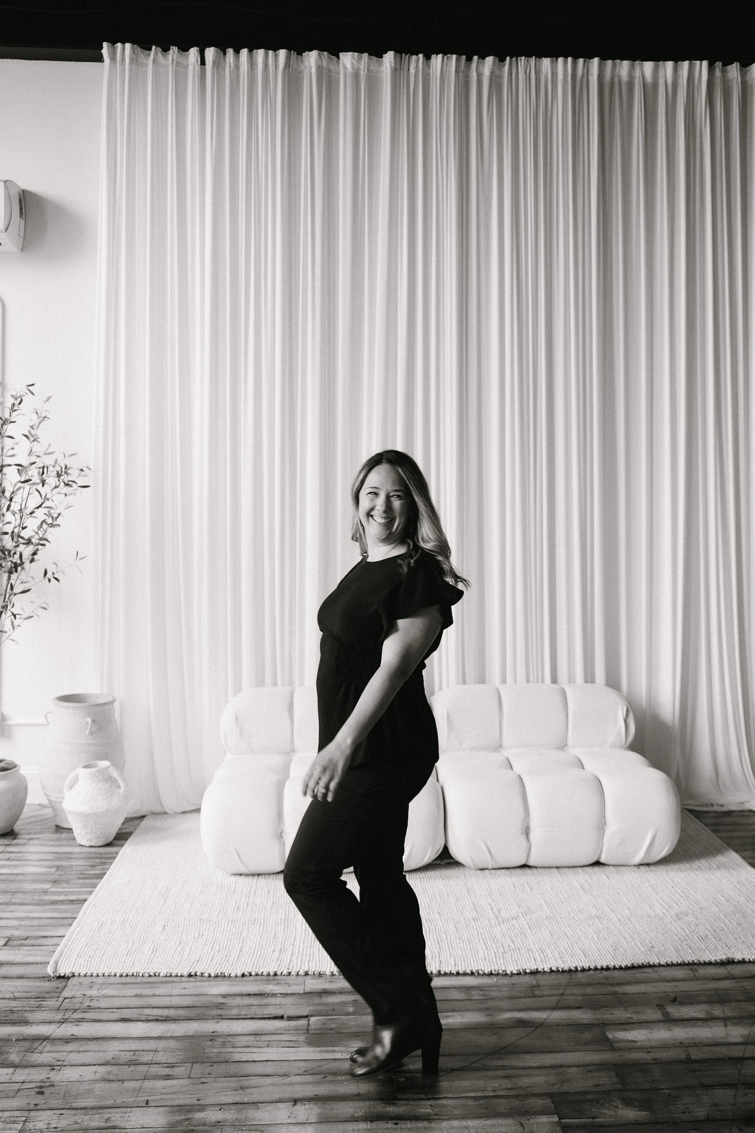 A woman with long hair smiling and standing in a living room with a white couch, curtains, potted plant, and vases, dressed in a black dress and high heels.