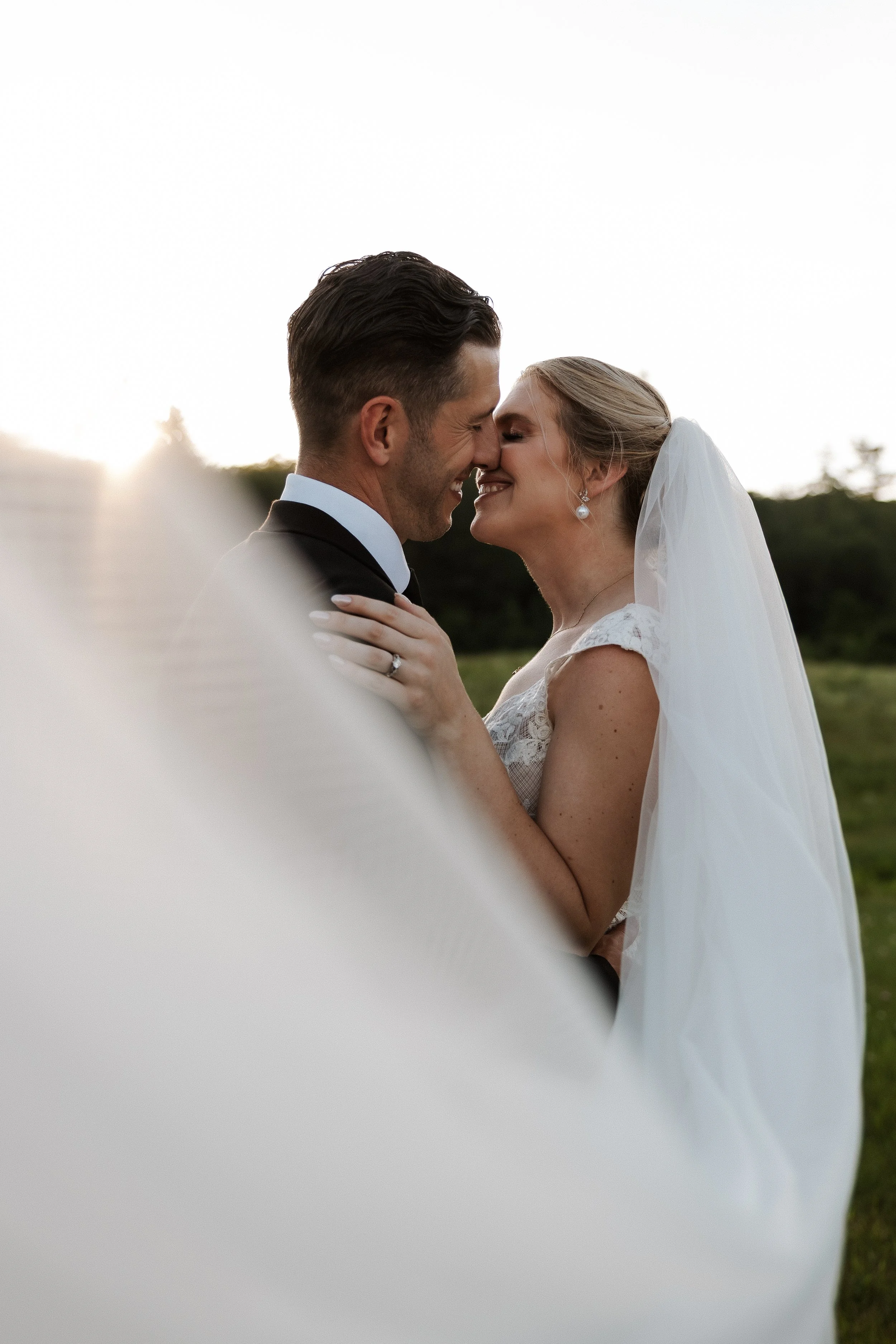 A bride and groom standing close outdoors, touching noses and smiling with their eyes closed, during sunset or late afternoon, with trees and sky in the background.
