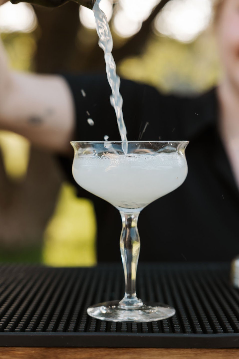 A cocktail glass with a cloudy, white beverage being poured into it on a black bar mat, with a person in black and yellow in the background.
