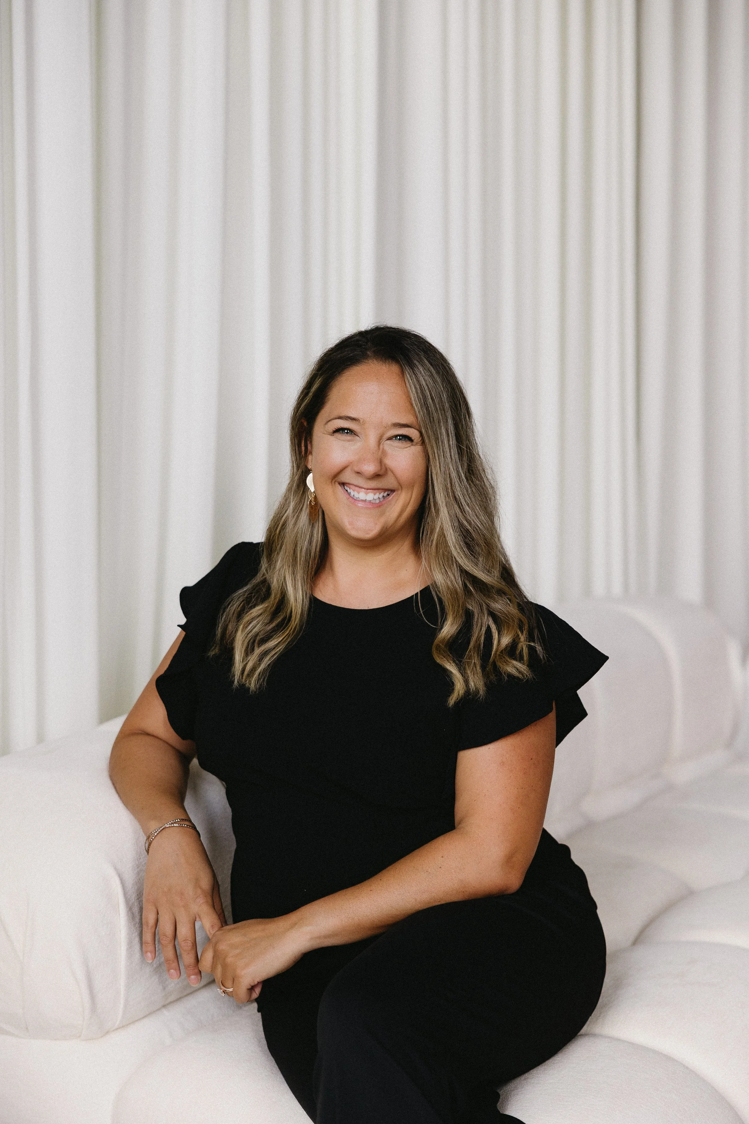 A woman with long blonde hair, wearing a black dress, smiling as she sits on a white sofa in a room with white curtains.