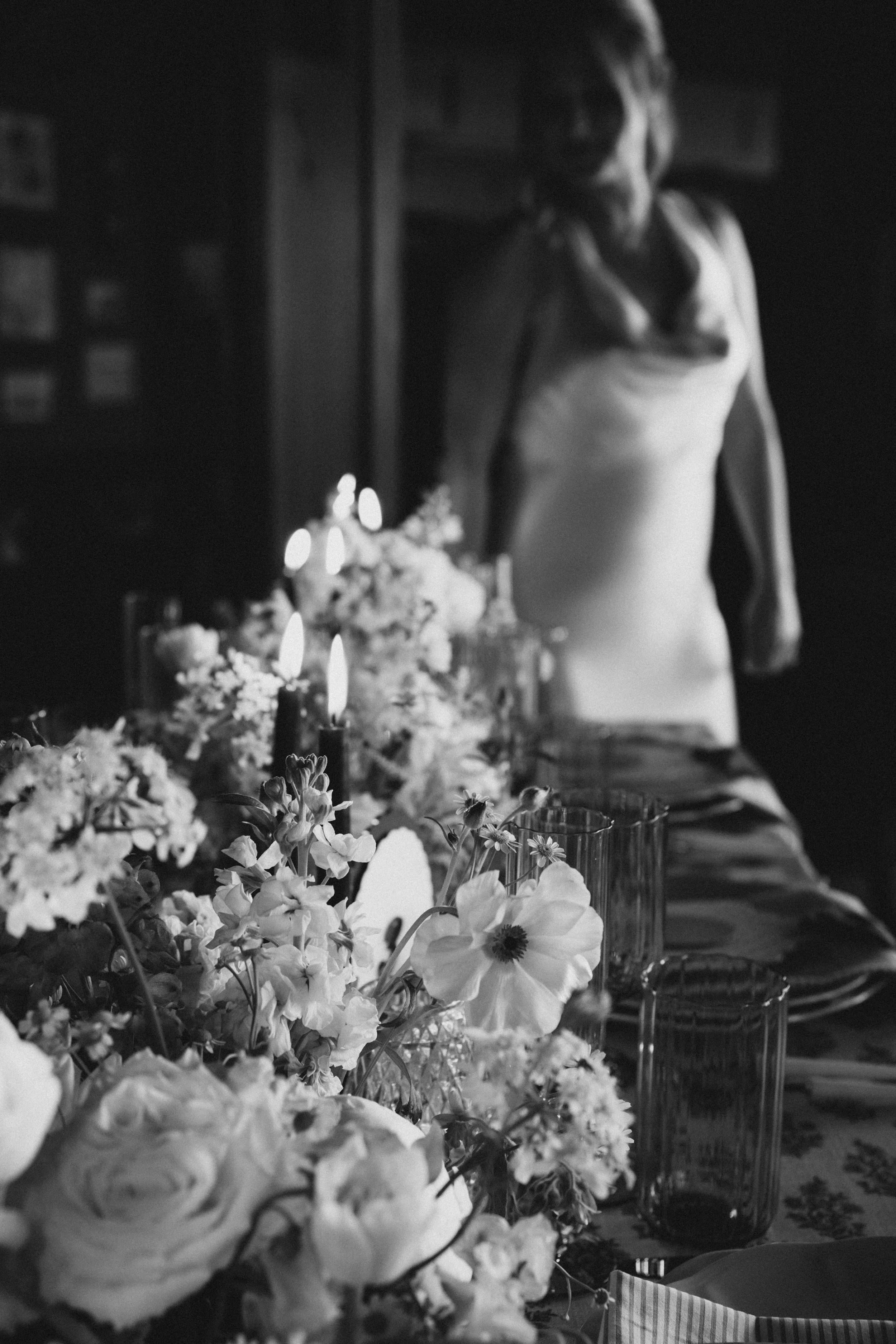 Black and white photo of a woman touching her face, surrounded by flowers and candles on a table.