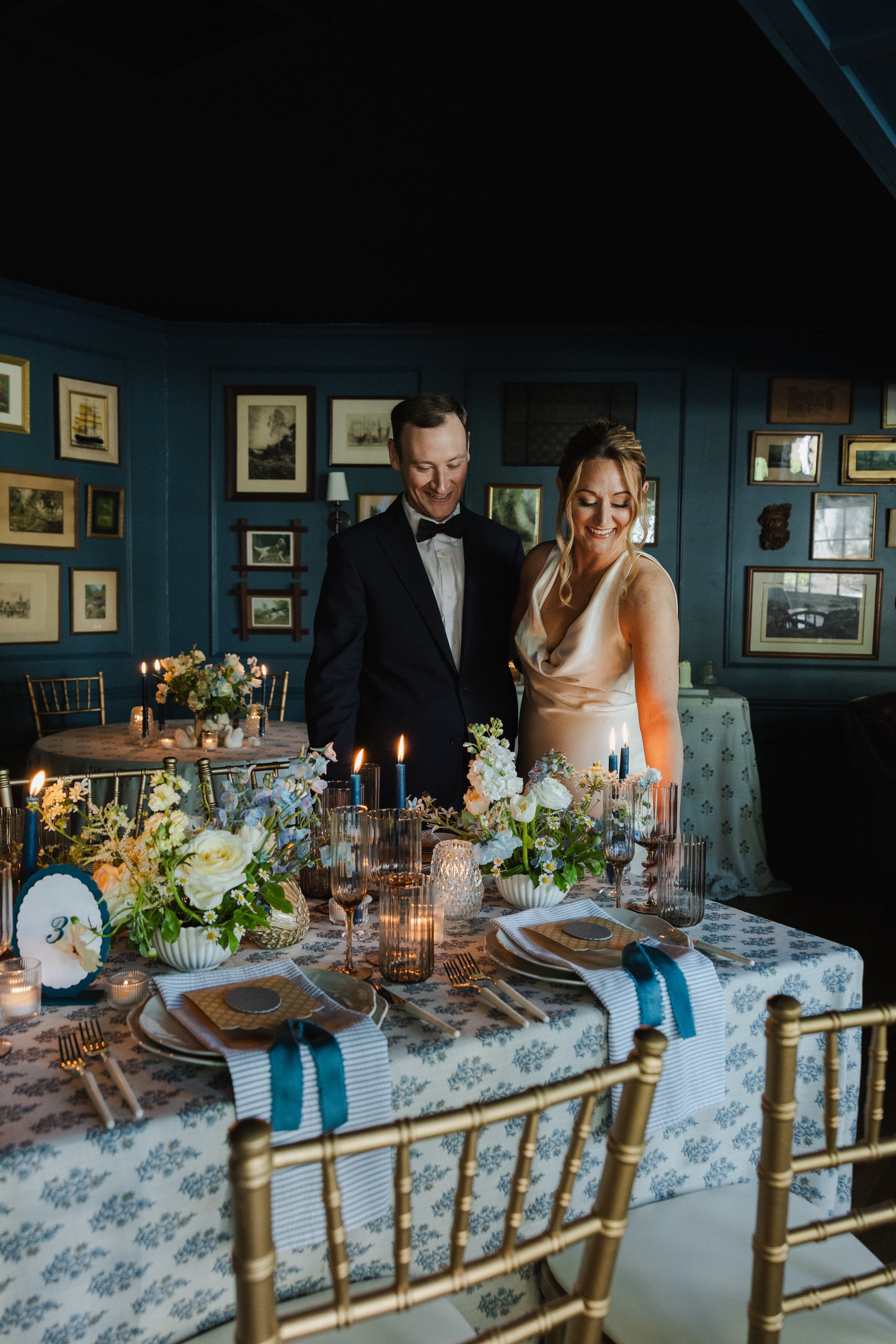 A wedding reception with a long decorated table, floral centerpieces, candles, and placed dinnerware, where a bride and groom are standing and smiling.
