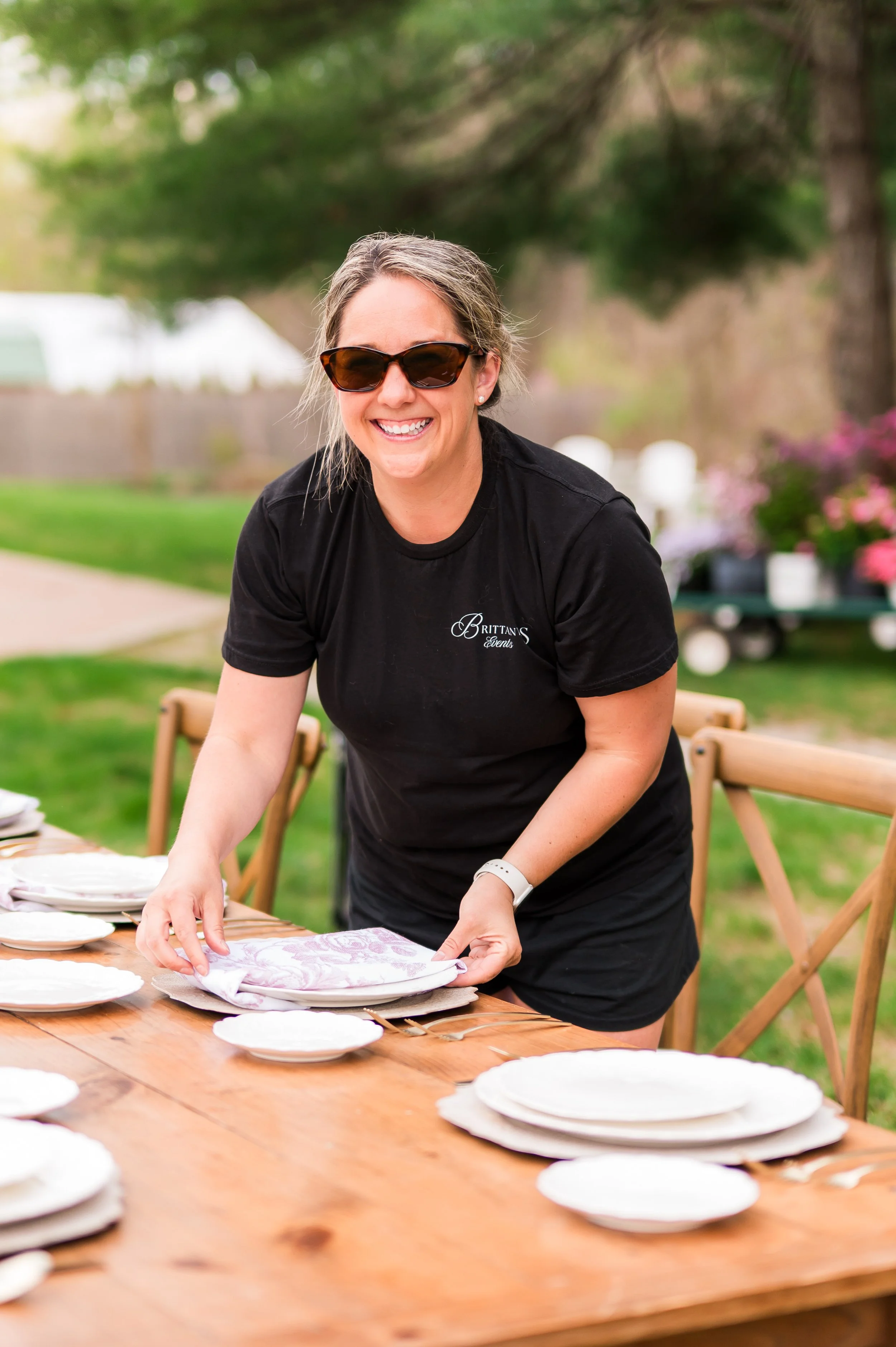 A woman with sunglasses and a black t-shirt is setting a table outdoors, with chairs and a dessert table in the background.