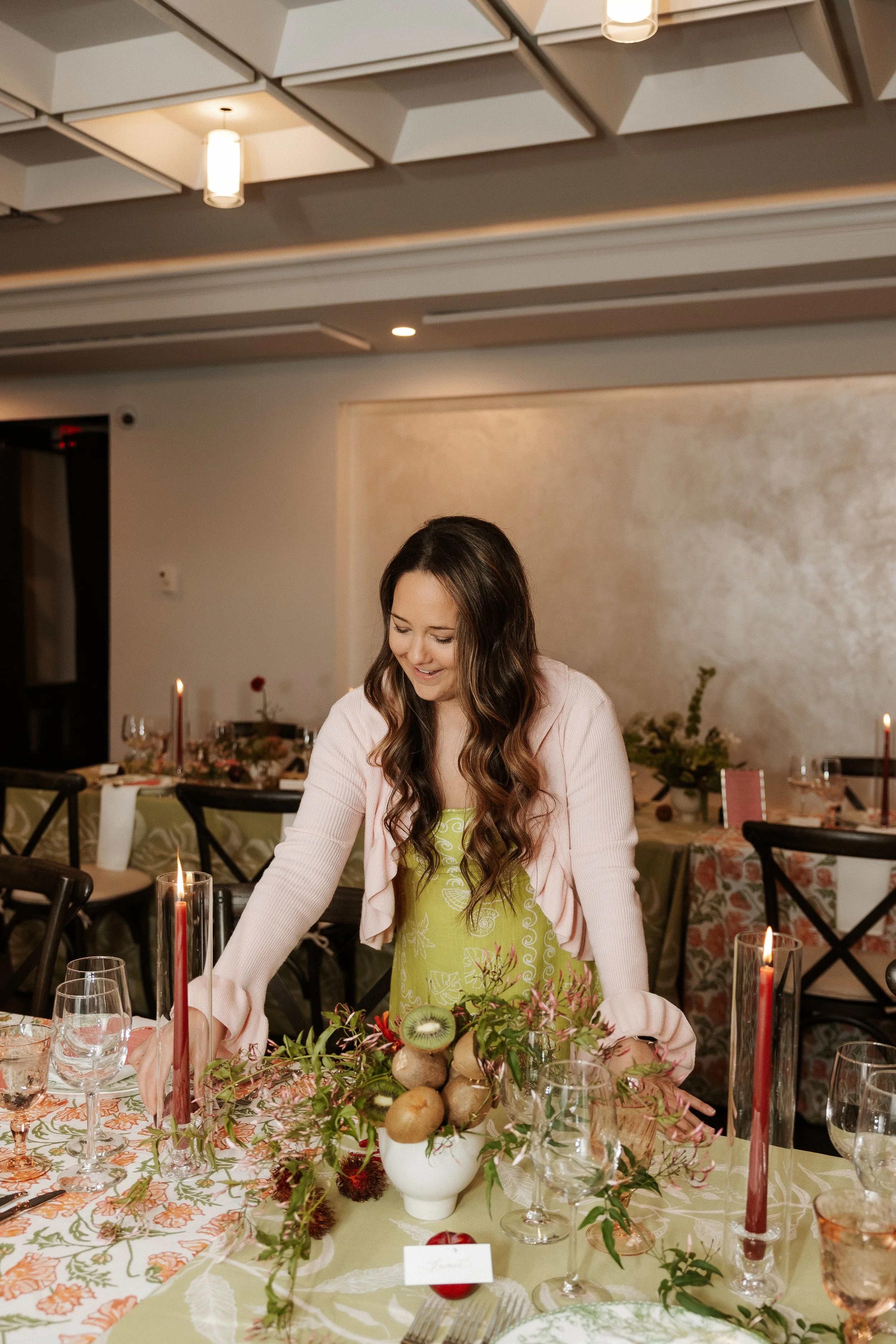 A woman with long curly hair wearing a pink cardigan and a green dress with white floral patterns standing at a decorated dining table in a restaurant. The table has a floral tablecloth, a centerpiece with fruits and foliage, and candles. The backgro