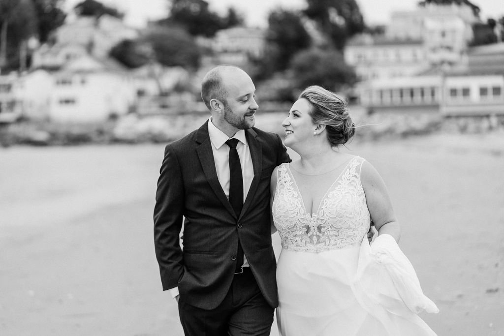Black and white photo of a bride and groom walking on the beach, looking at each other and smiling. The groom wears a suit and tie, and the bride wears a wedding dress with lace details.