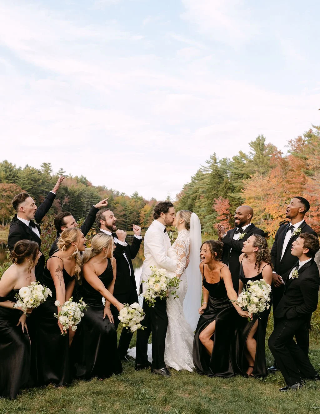 A wedding party outdoors with a bride and groom in the center, surrounded by bridesmaids and groomsmen, all celebrating and smiling, with trees and autumn foliage in the background.