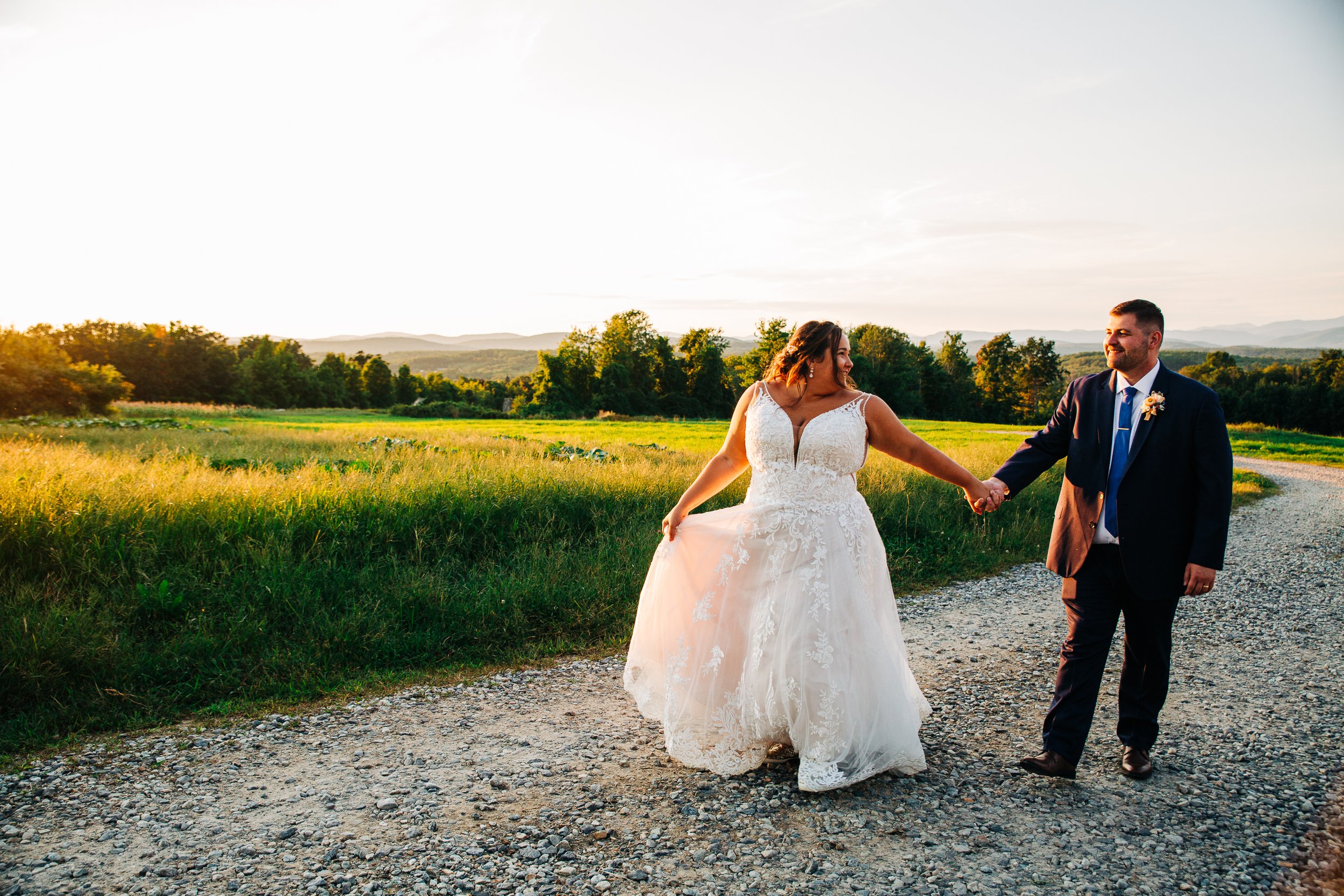 Bride and groom holding hands and walking on a gravel path in a rural landscape at sunset, with green fields and trees in the background.