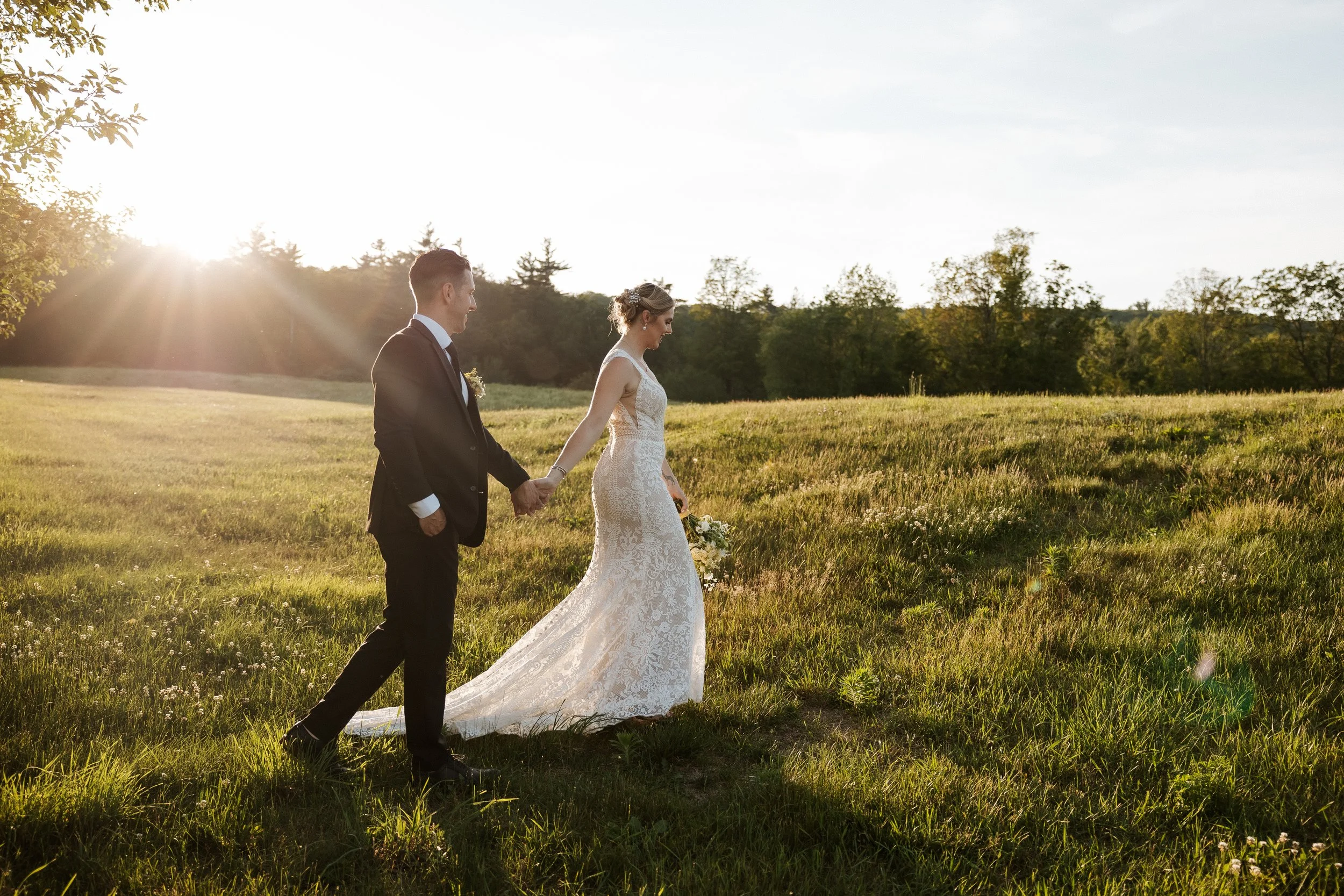 A bride and groom holding hands and walking through a grassy field during sunset, with trees in the background.
