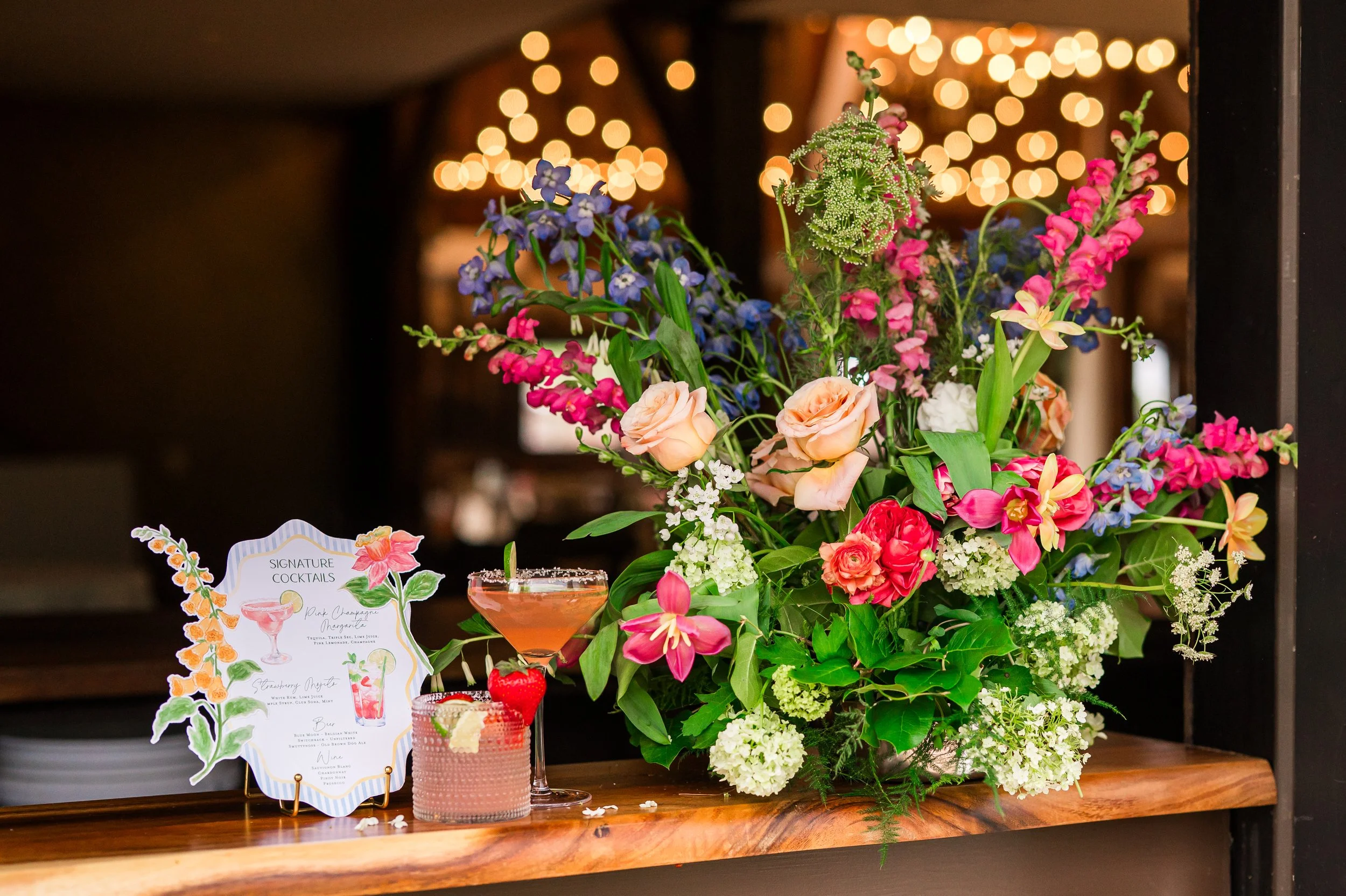 A vibrant floral arrangement with pink, purple, and white flowers sits on a wooden table, decorated with a cocktail, a paper menu, and a sign labeled 'Signature Cocktails' with illustrations of drinks and flowers, against a background of warm, blurred string lights.