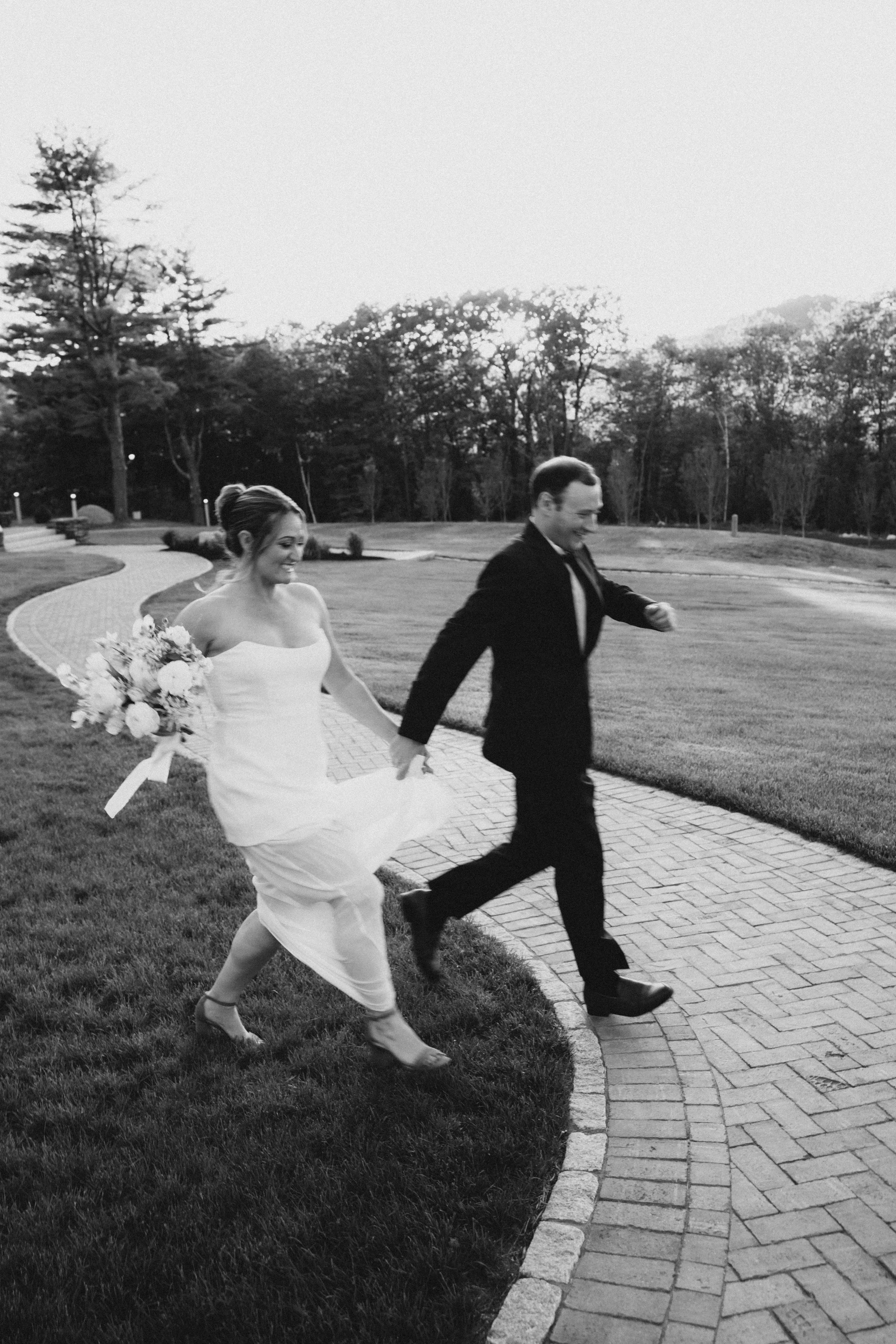 A black-and-white photo of a bride and groom holding hands and running along a winding brick path in a park, with trees and a grassy lawn in the background.