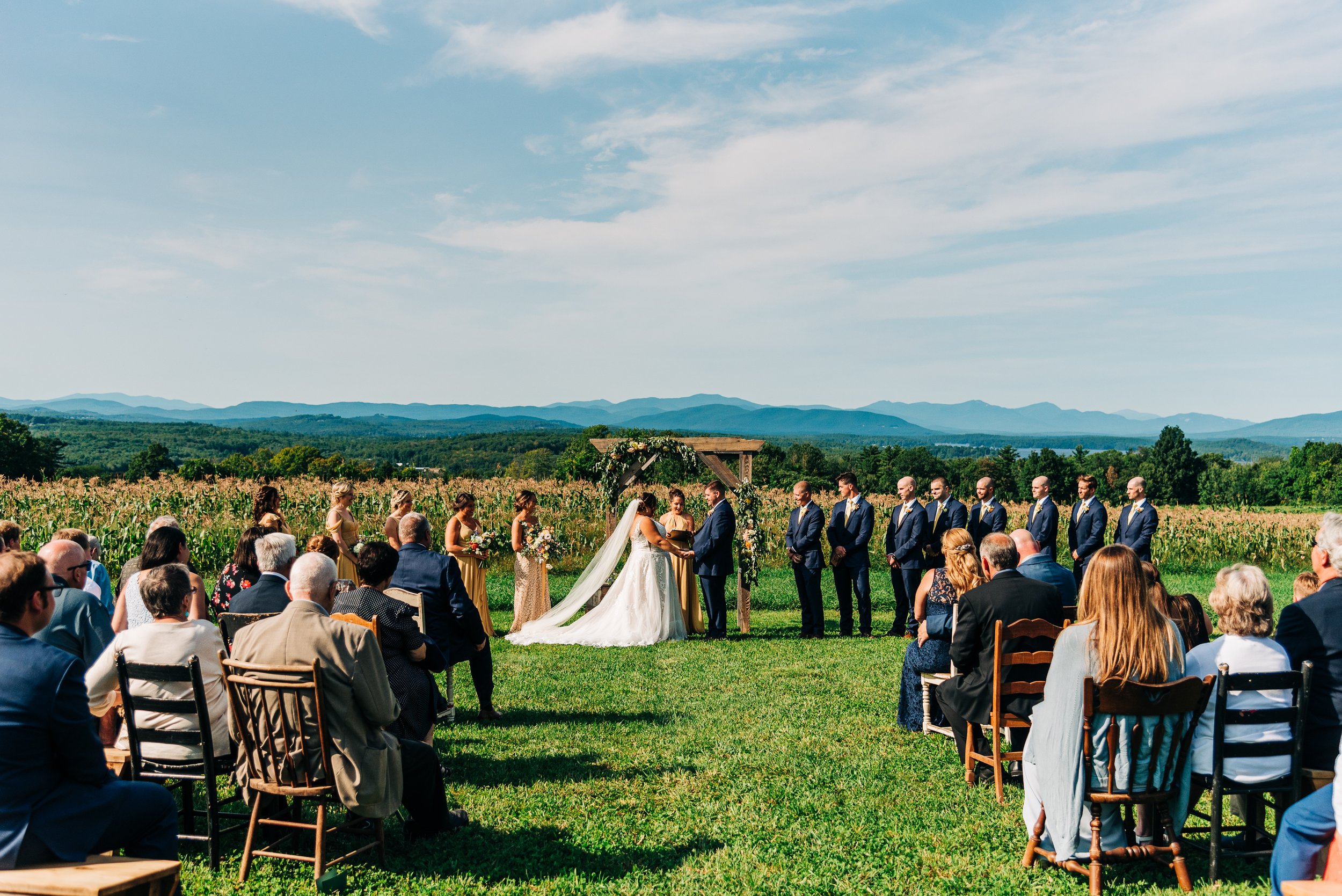 Outdoor wedding ceremony in a field with a seated guests, a bride and groom in front of a wooden arch, and a backdrop of mountains and a blue sky.
