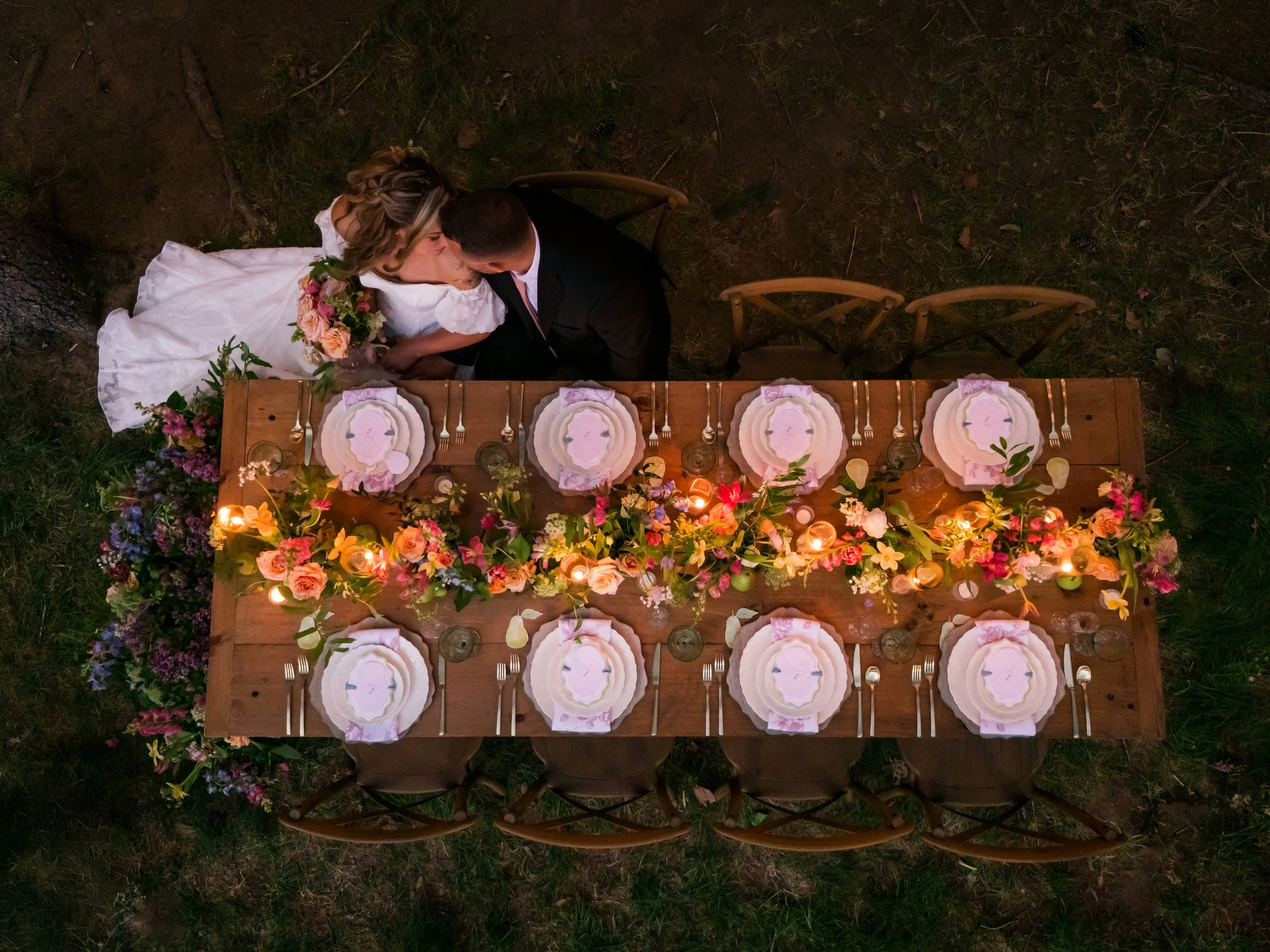 Overhead shot of a bride and groom kissing at a wedding reception table decorated with flowers, candles, and tableware on a grassy outdoor area.