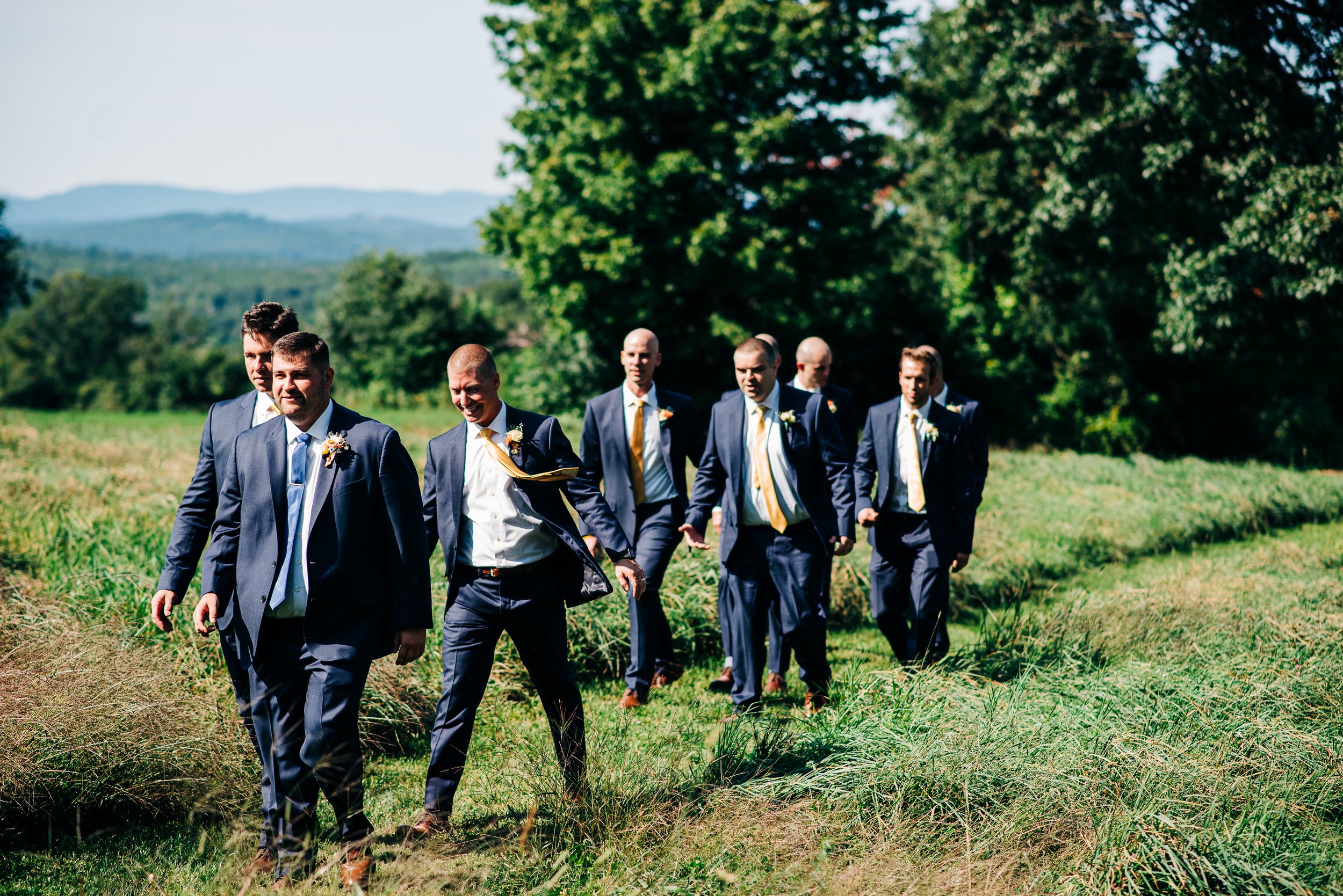 A group of men dressed in suits walking outdoors in a green field with trees and mountains in the background.