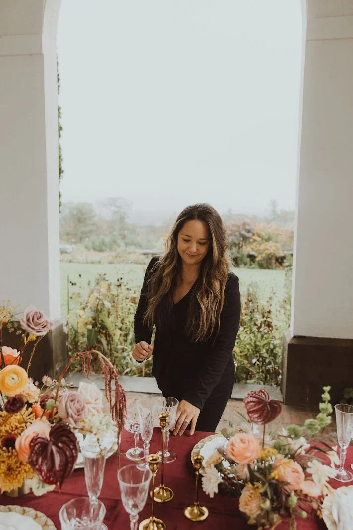 A woman with long brown hair, wearing a black outfit, stands behind a decorated table with flowers and glassware, inside a room with an open view of a garden or field outside.